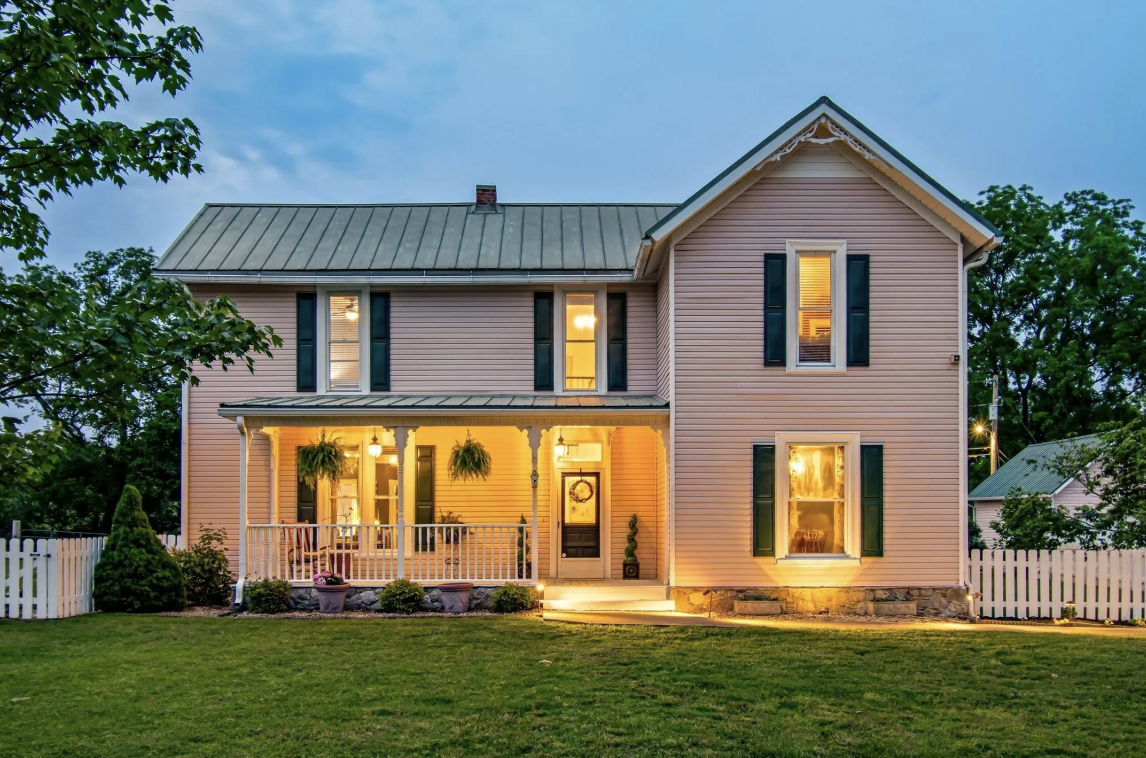 The image shows a two-story, pastel pink house with a green metal roof, white trim, and a front porch, surrounded by greenery and a white picket fence.