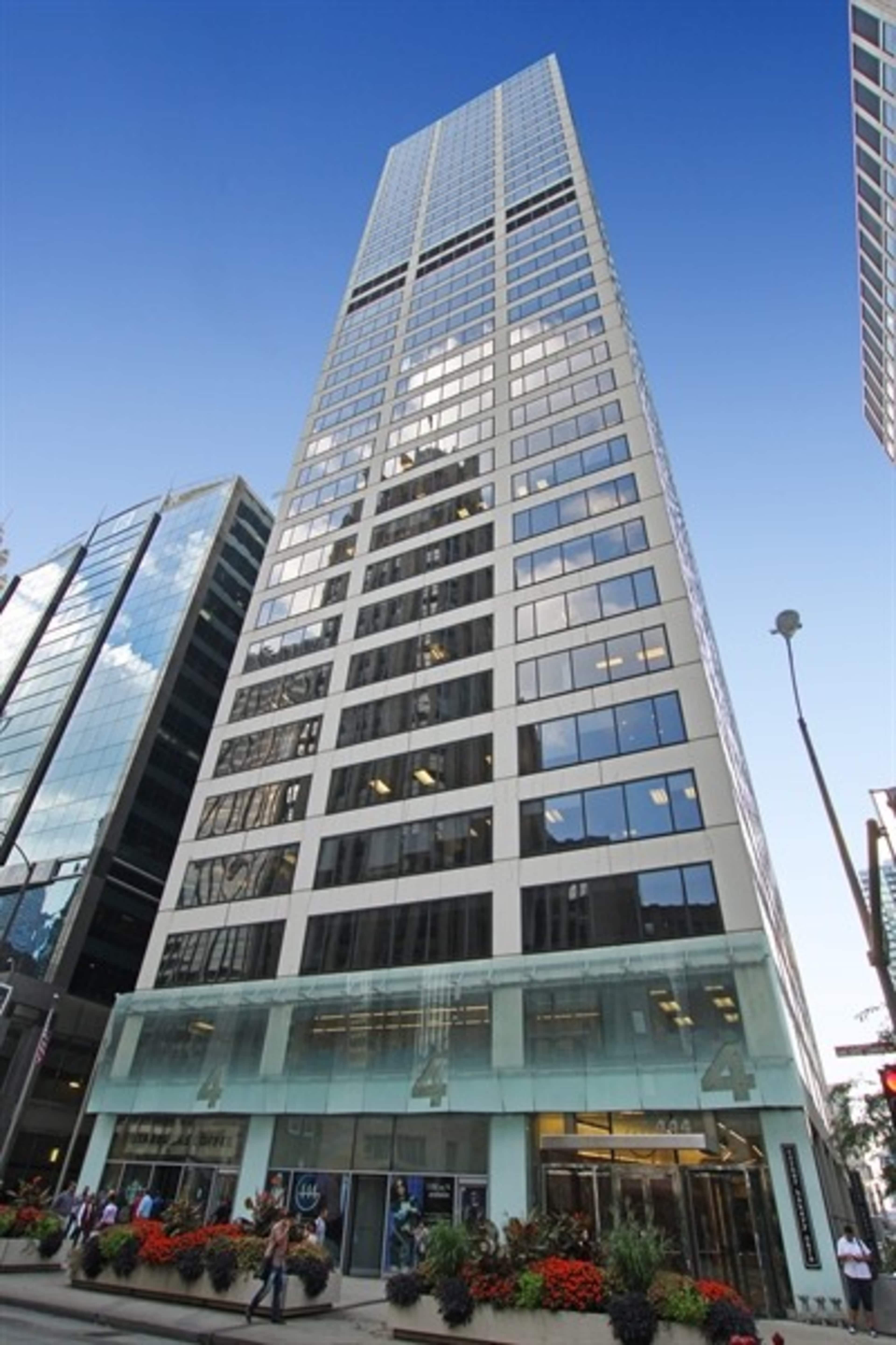 A tall, modern office building with a glass facade is situated among other skyscrapers, featuring a transparent entrance and colorful flower beds in front.