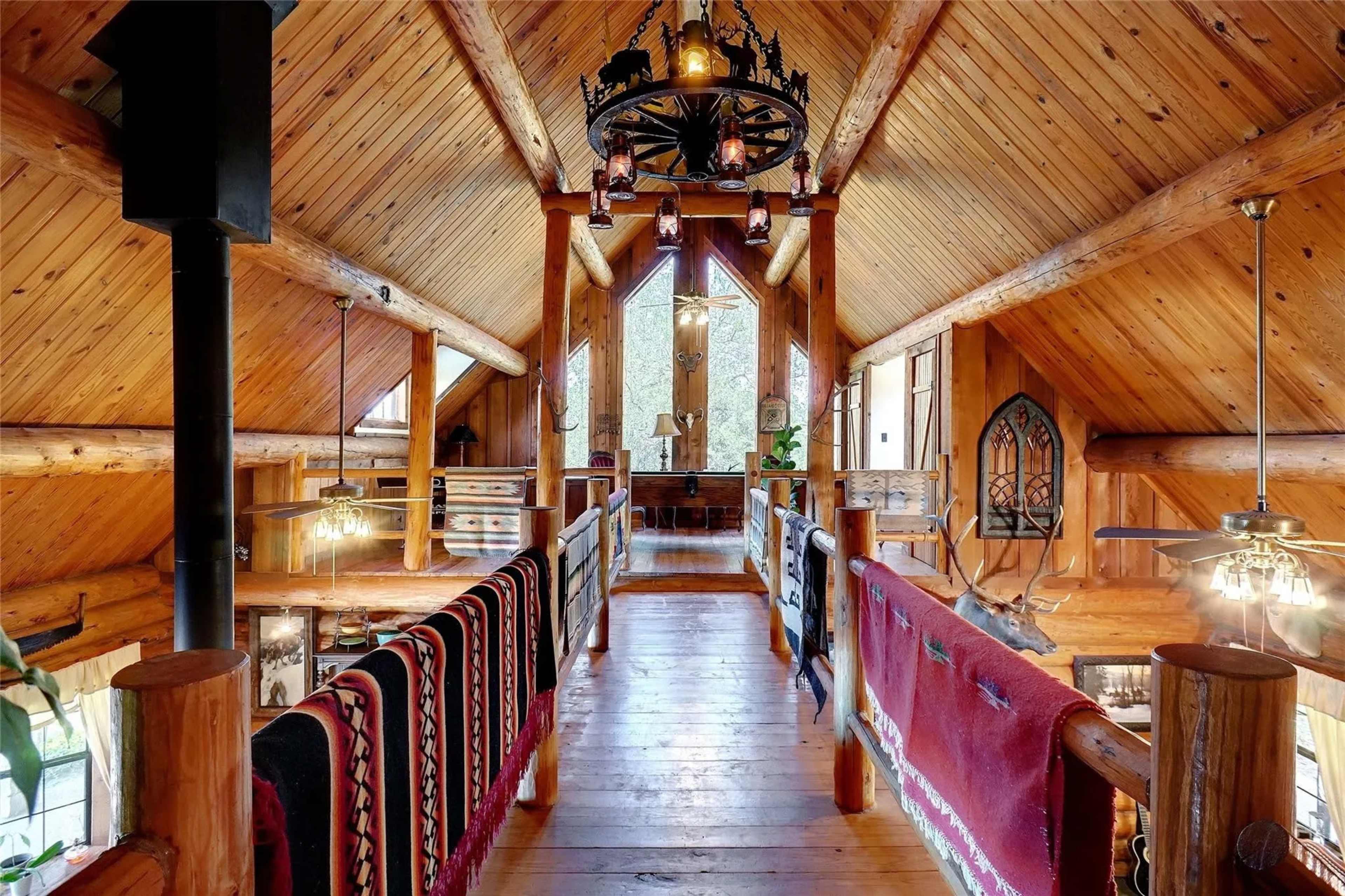 A rustic wooden loft with exposed beams, a high ceiling, and a balcony overlooking a spacious area below, adorned with decorative items and rugs.