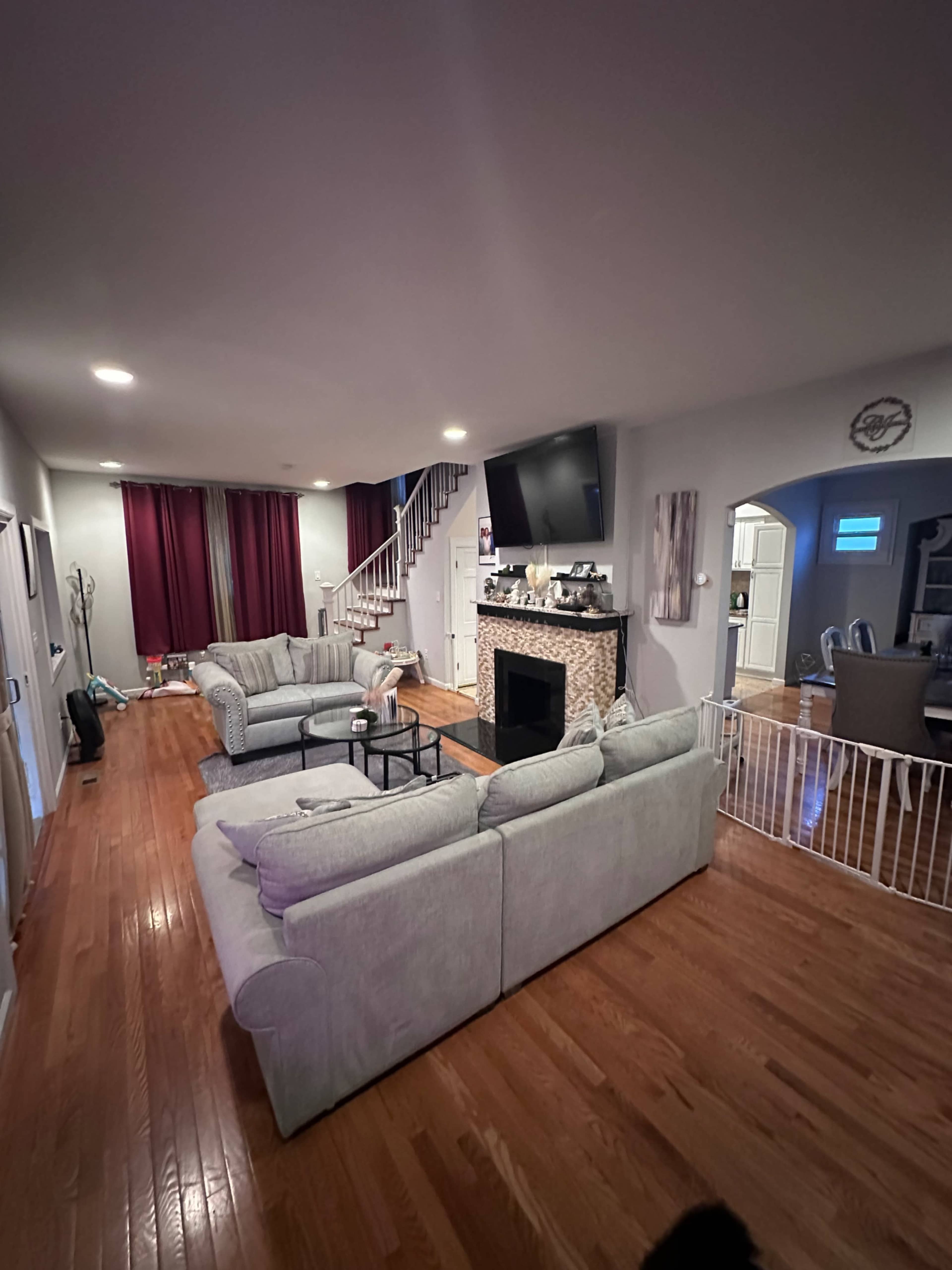 The image shows a spacious living room with gray sofas, a fireplace, a large television mounted on the wall, and a staircase leading upstairs.