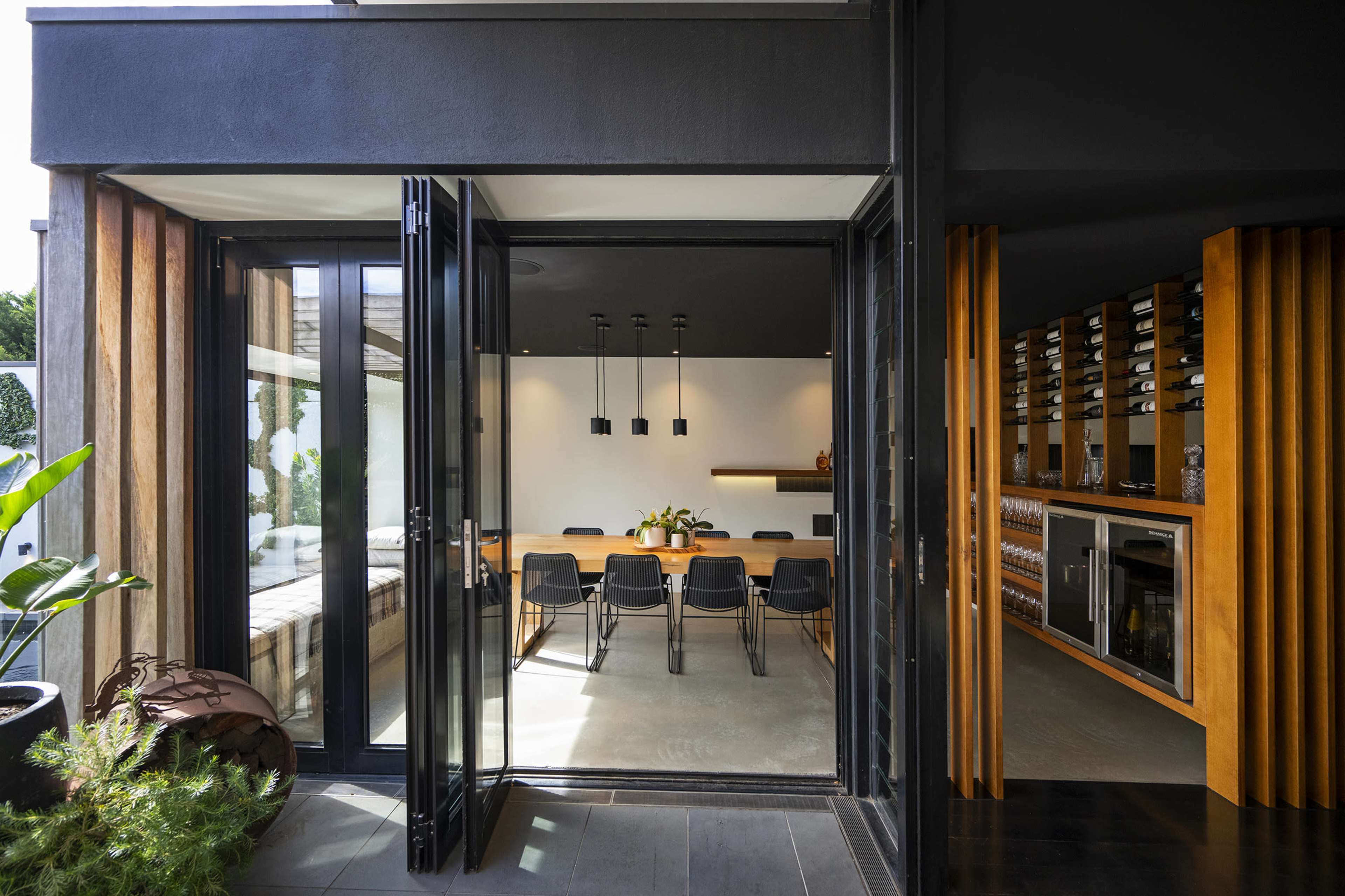 The image shows a modern dining area with a large wooden table and black chairs, adjacent to a wine storage space, with glass doors leading to an outdoor area.
