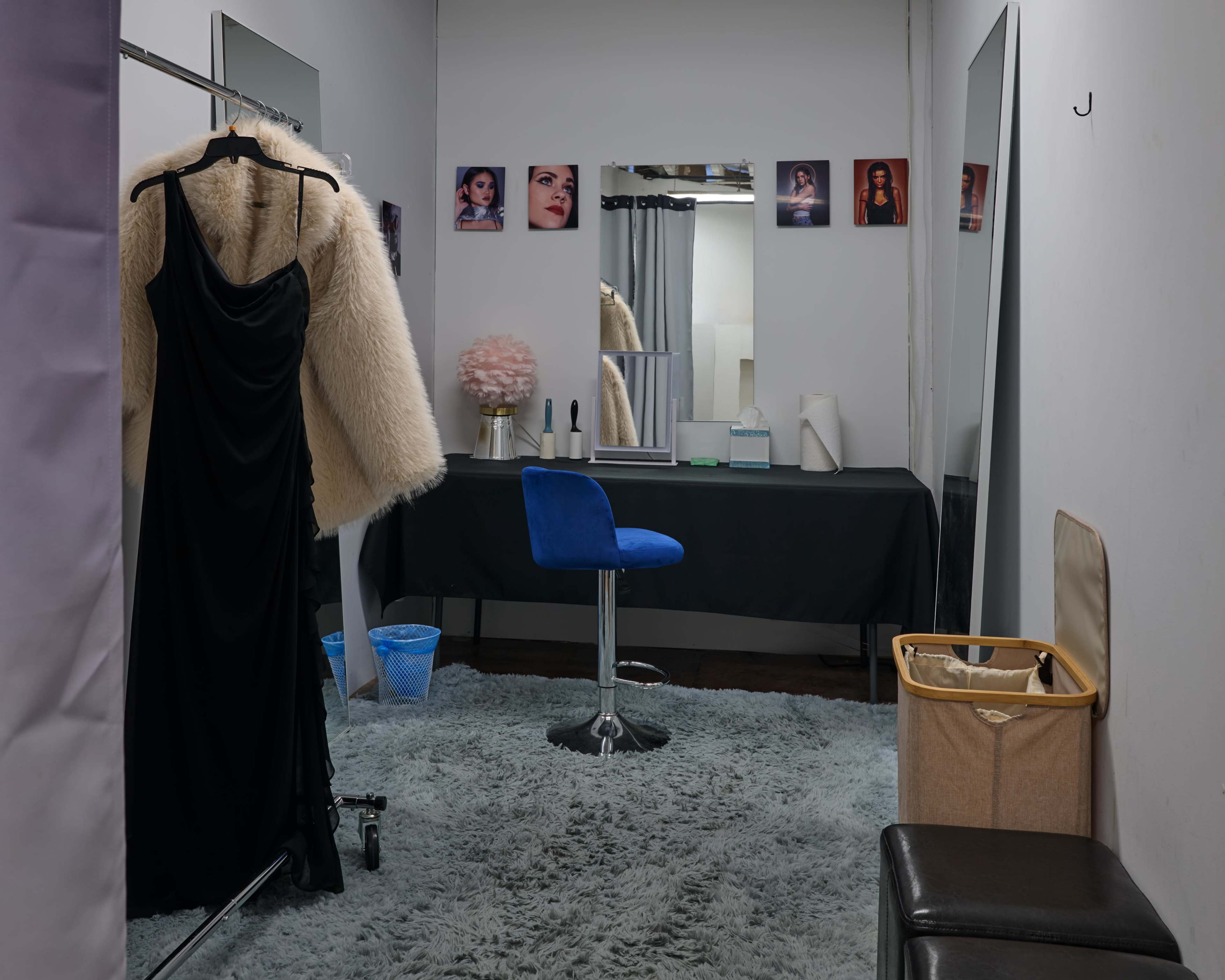 The image shows a small dressing room with a black table, a blue chair, a mirror, and a black evening gown hung on a rack, complemented by portraits on the wall and a plush rug on the floor.