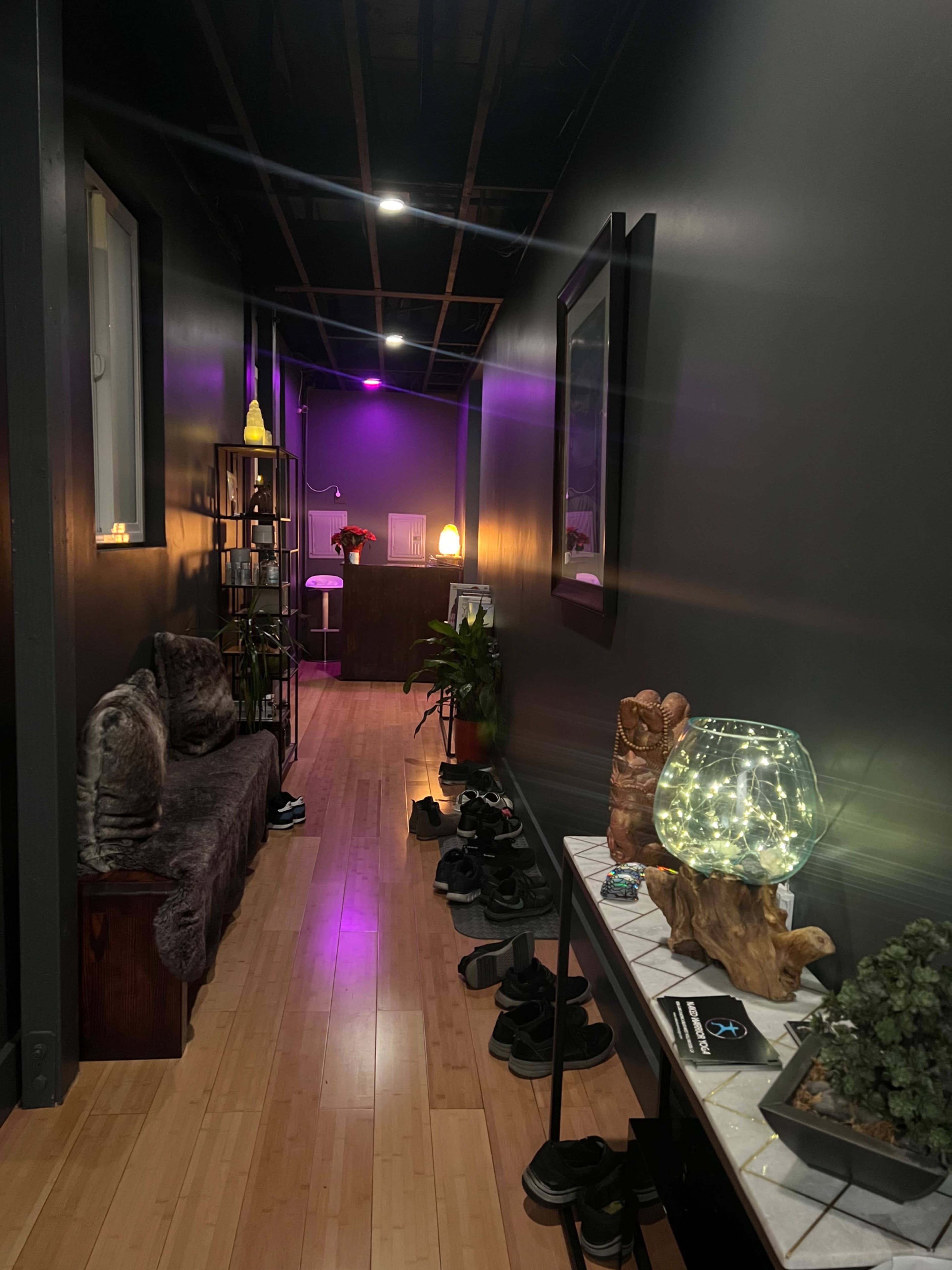 A dimly lit hallway with dark walls features a cozy seating area on one side, a shelf filled with decorative items, and shoes scattered on the floor.
