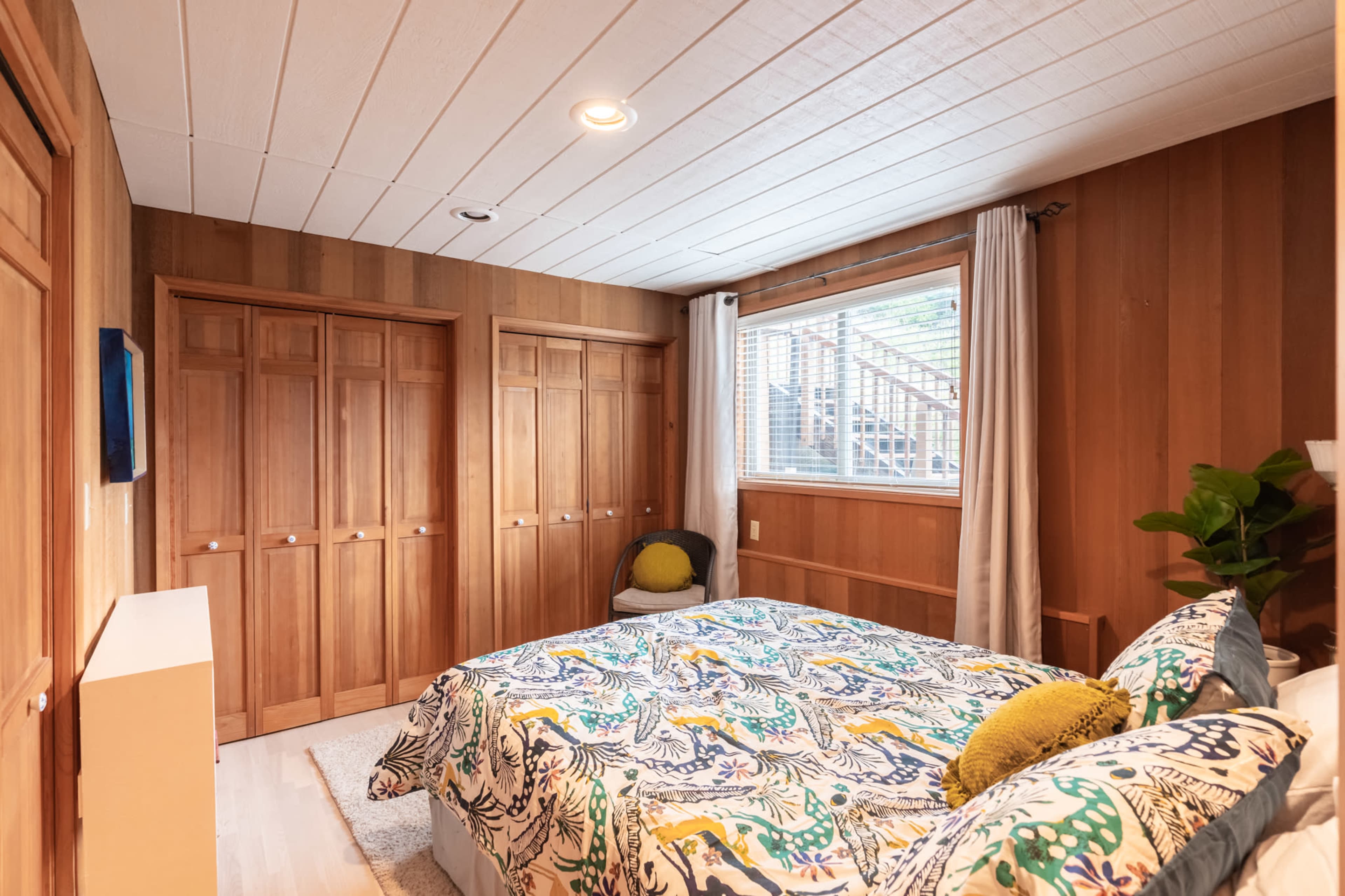 The image shows a wooden-paneled bedroom with a bed adorned in a colorful floral-patterned duvet and a window revealing an outdoor staircase.