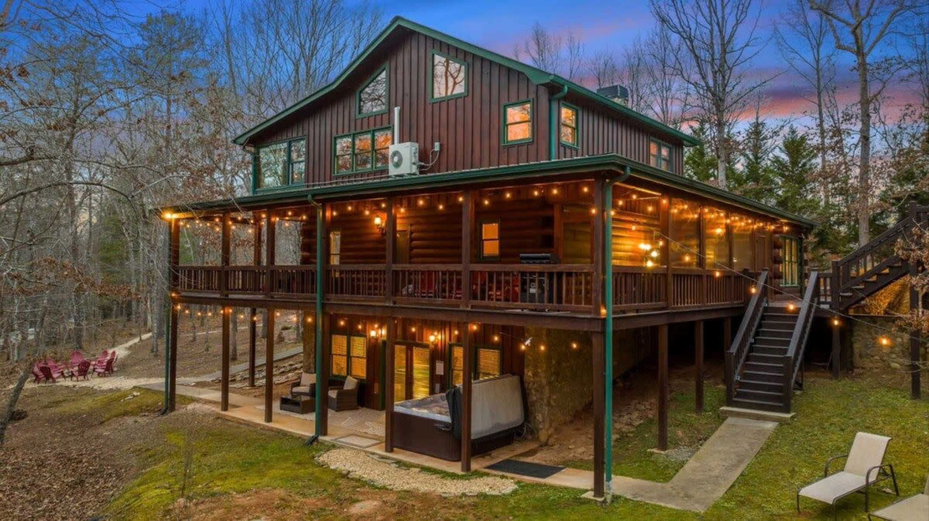 A large wooden cabin with multiple levels and string lights is surrounded by trees in a wooded area.
