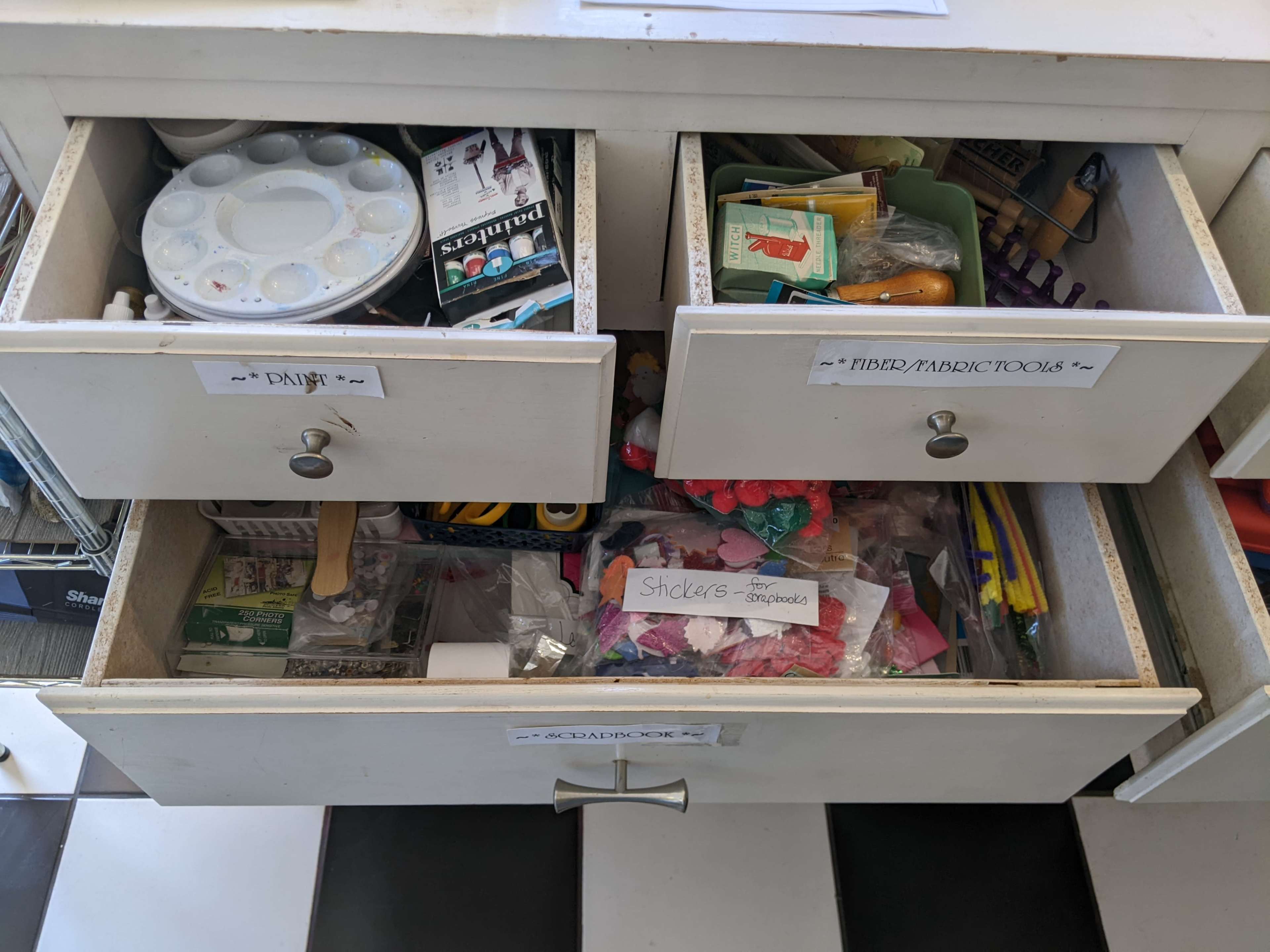 The image shows a cluttered drawer with art supplies, including palettes, paint tubes, and various craft materials, organized into labeled compartments.