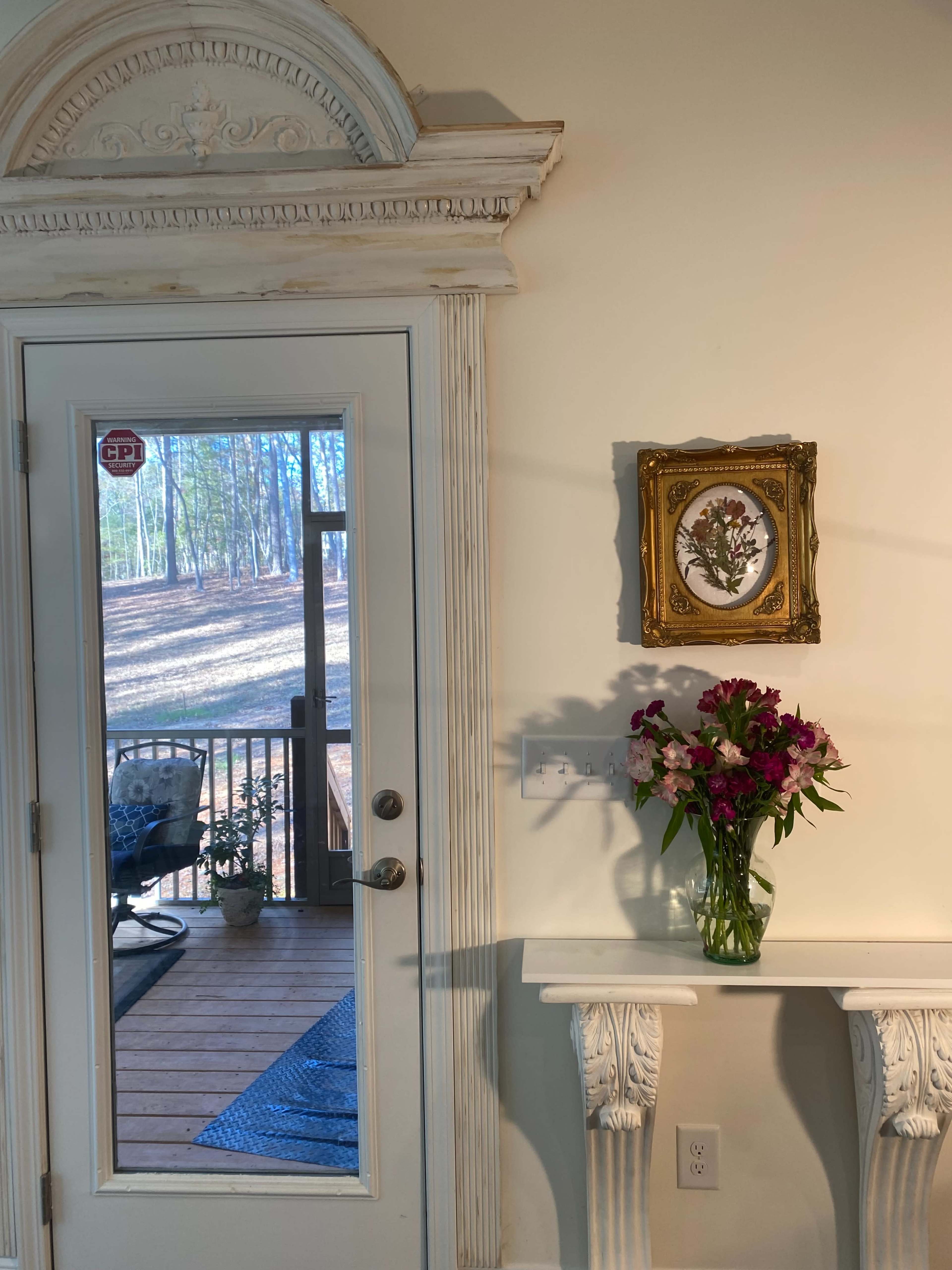 A framed painting hangs on a wall beside a vase of flowers, while a glass door leads to a wooden deck with a view of trees.