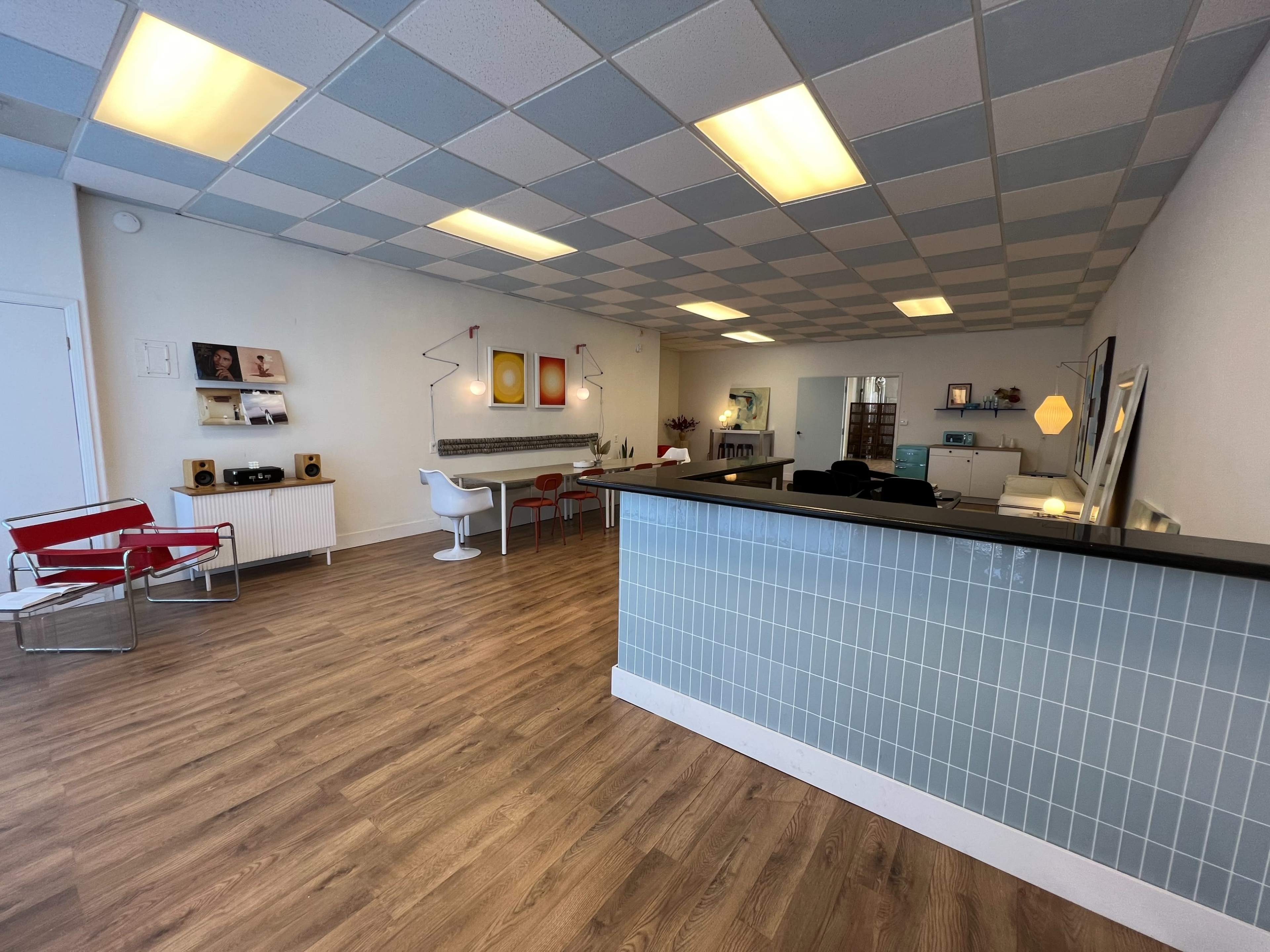 A modern interior space with a reception area, dining table, and seating arrangements, featuring a tiled counter and wooden floors.