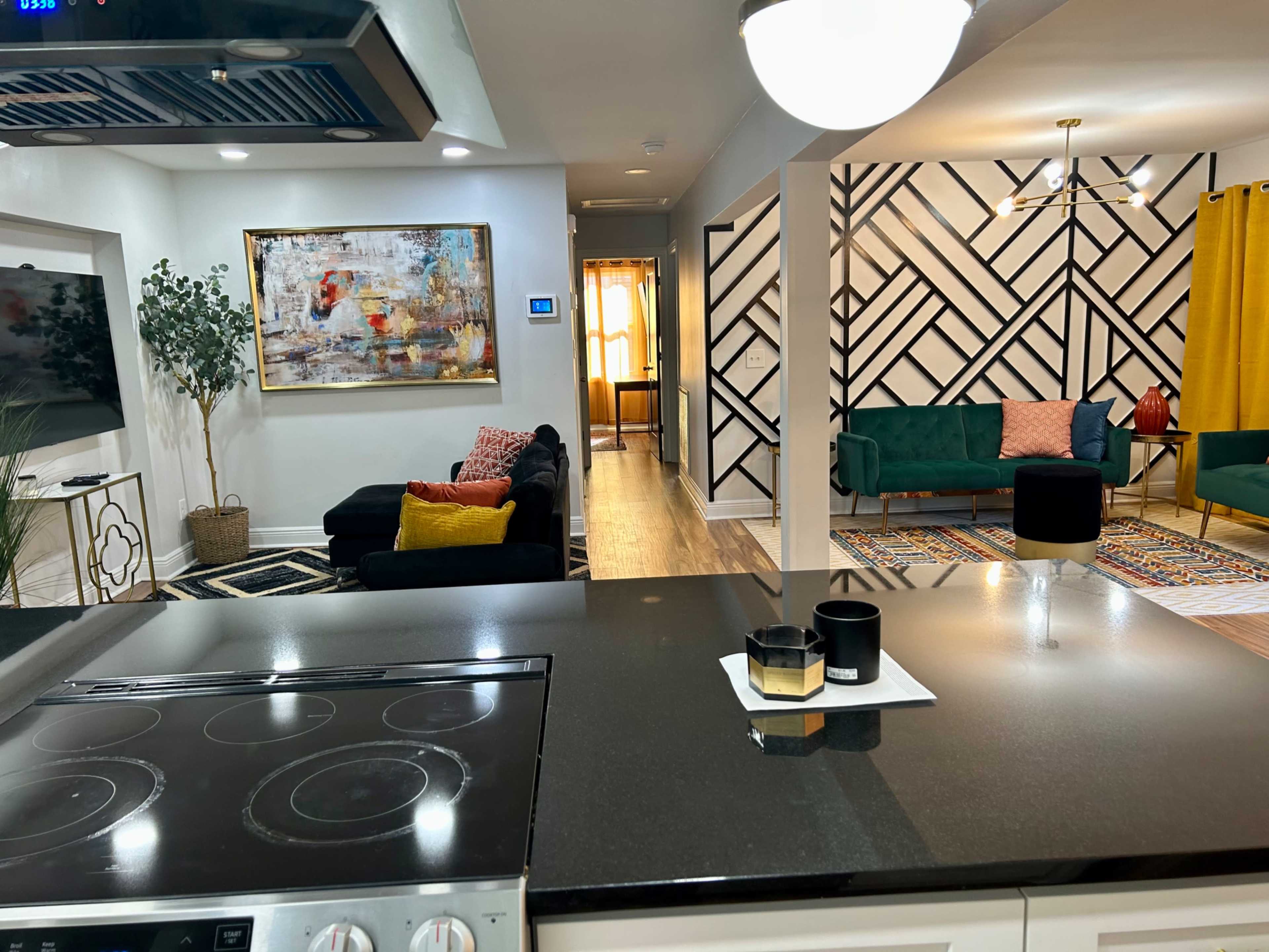 A modern open-concept living space featuring a kitchen with a dark countertop, a living area with a black sofa, and a brightly decorated room with green sofas and geometric wall patterns.