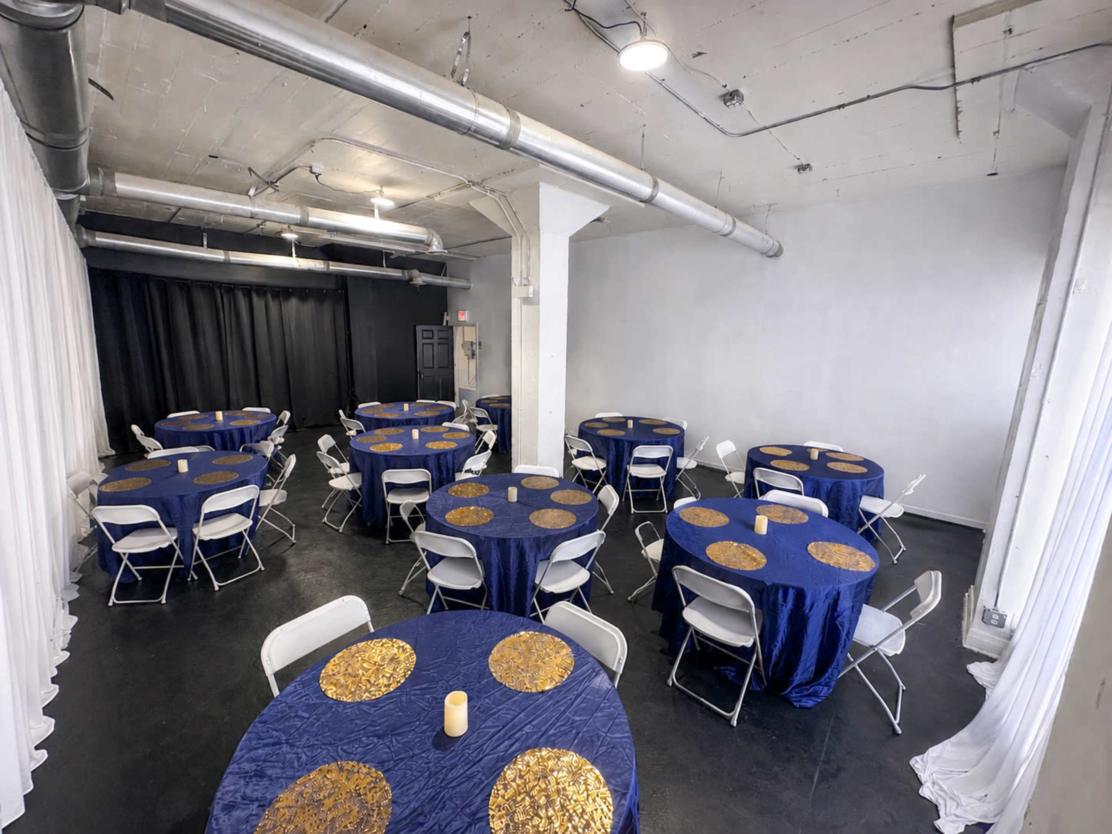 The image shows a spacious indoor venue with several round tables covered in blue tablecloths adorned with gold circular accents, arranged in a simple layout.