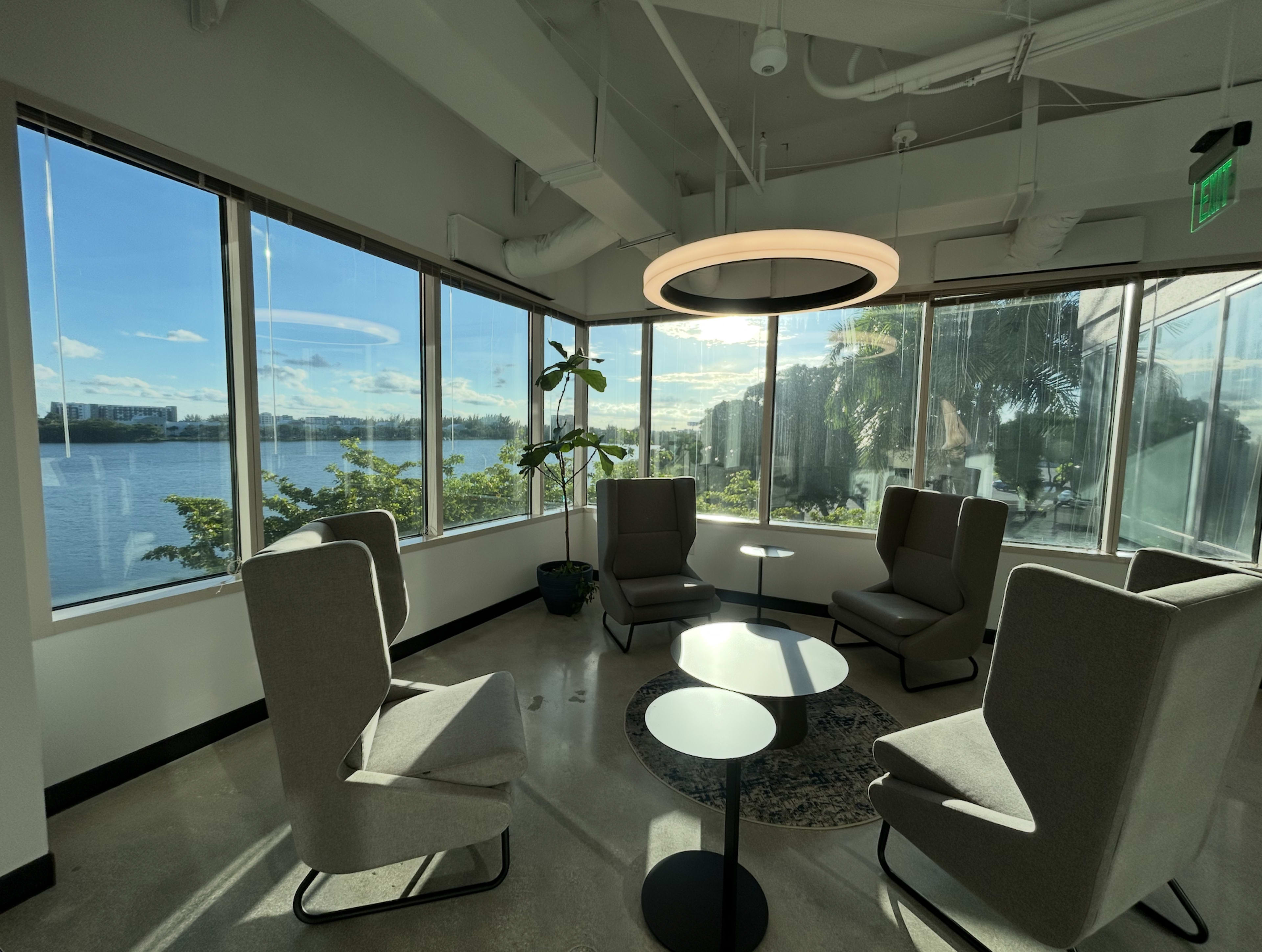 The image shows a modern lounge area with four gray chairs arranged around a small circular table, illuminated by natural light from large windows overlooking a waterfront scene.