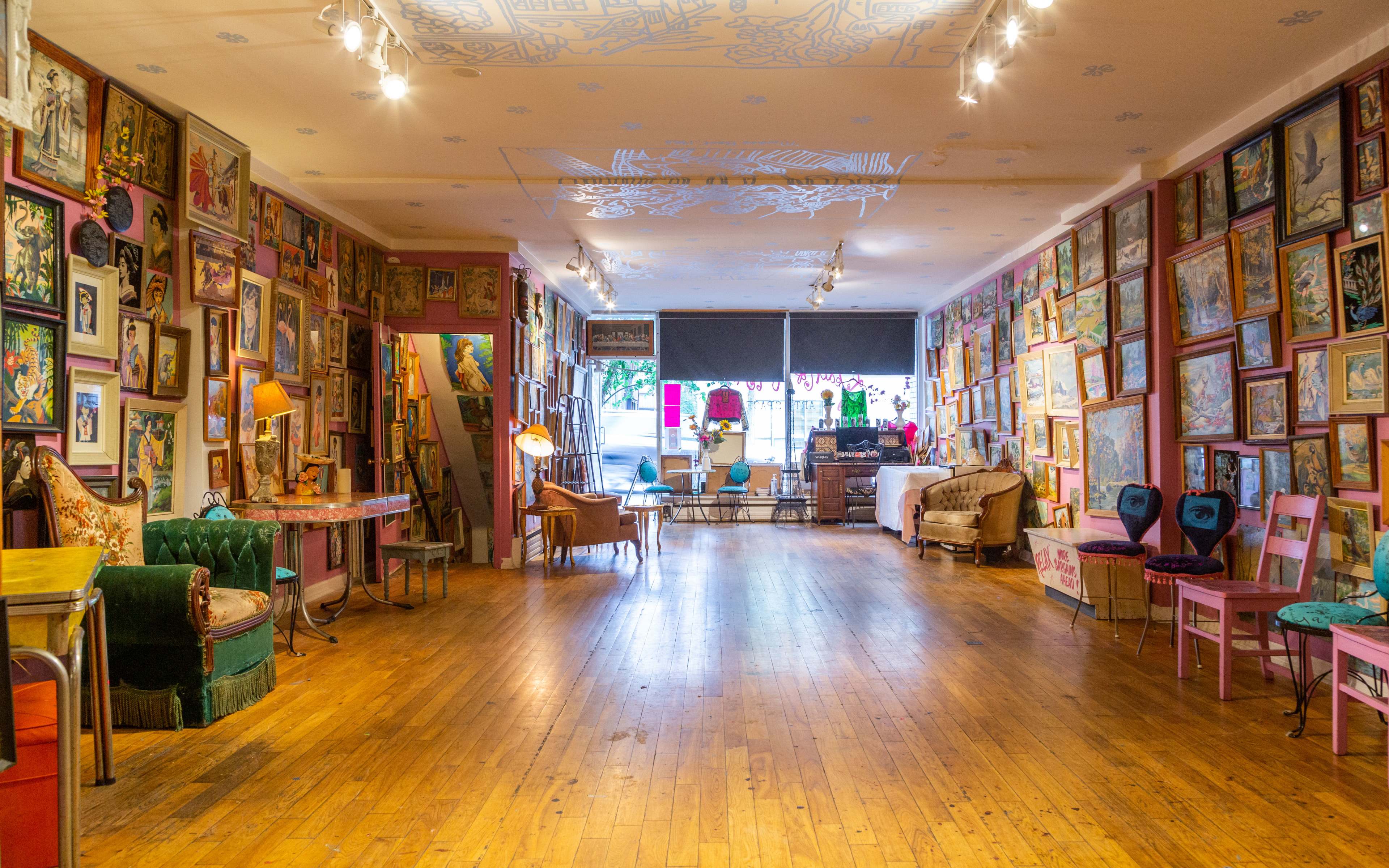 A spacious art gallery with walls covered in colorful framed artwork, wooden flooring, and a variety of furniture scattered throughout.