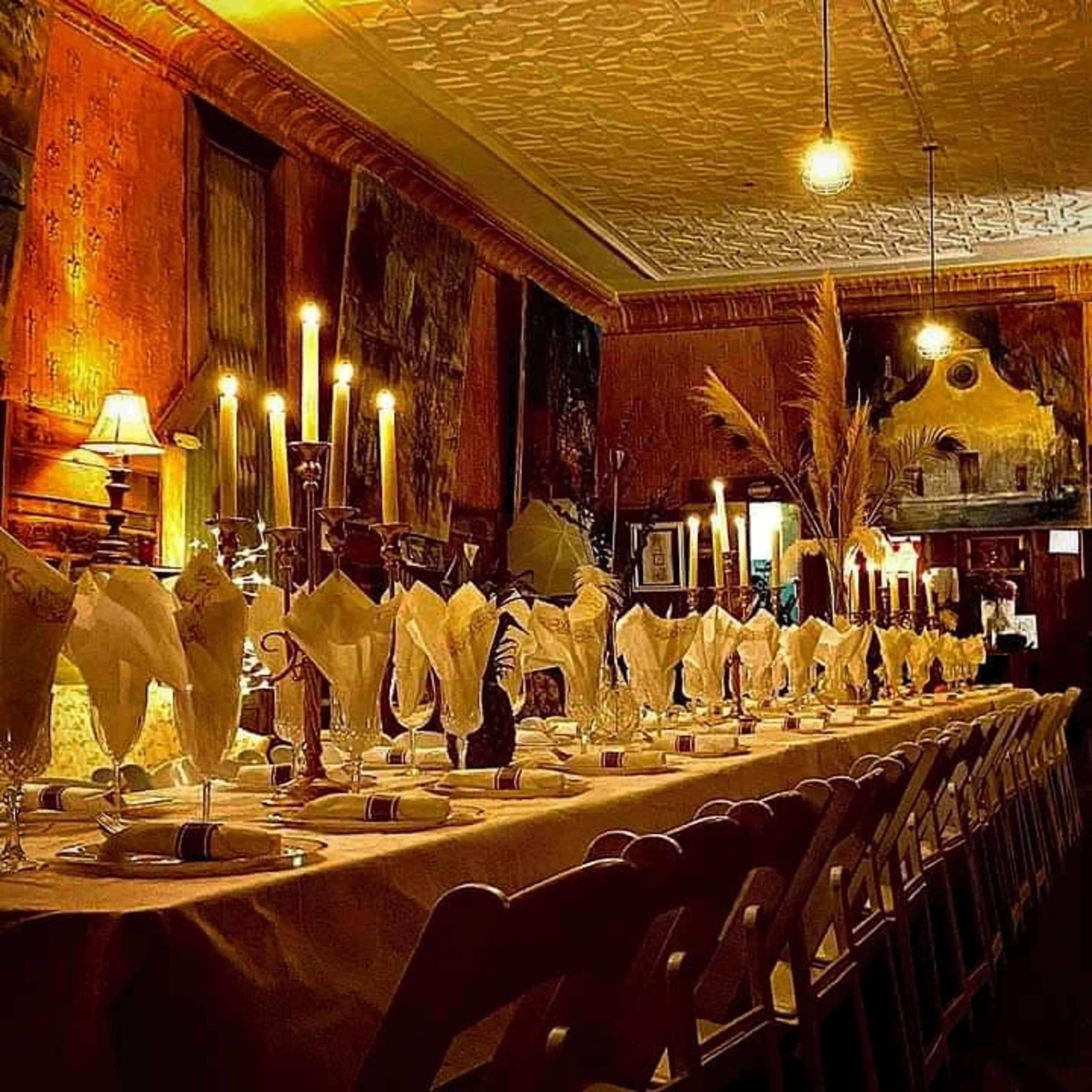 A long dining table is set with folded napkins, candles, and plates, illuminated by soft overhead lighting in a warmly decorated room.