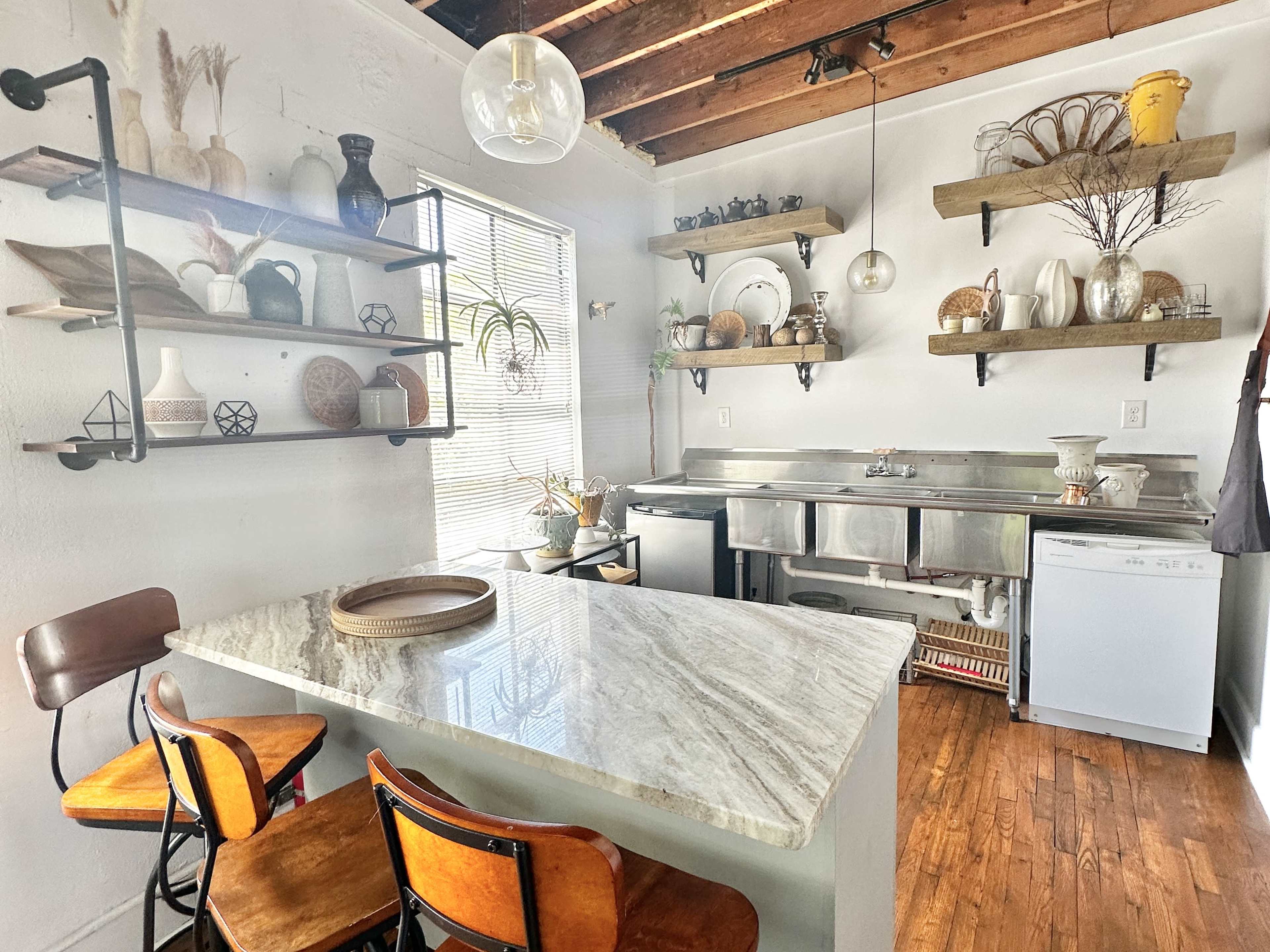 A modern kitchen features a granite countertop island with wooden bar stools, open shelves displaying various dishes and decorations, and stainless steel appliances.