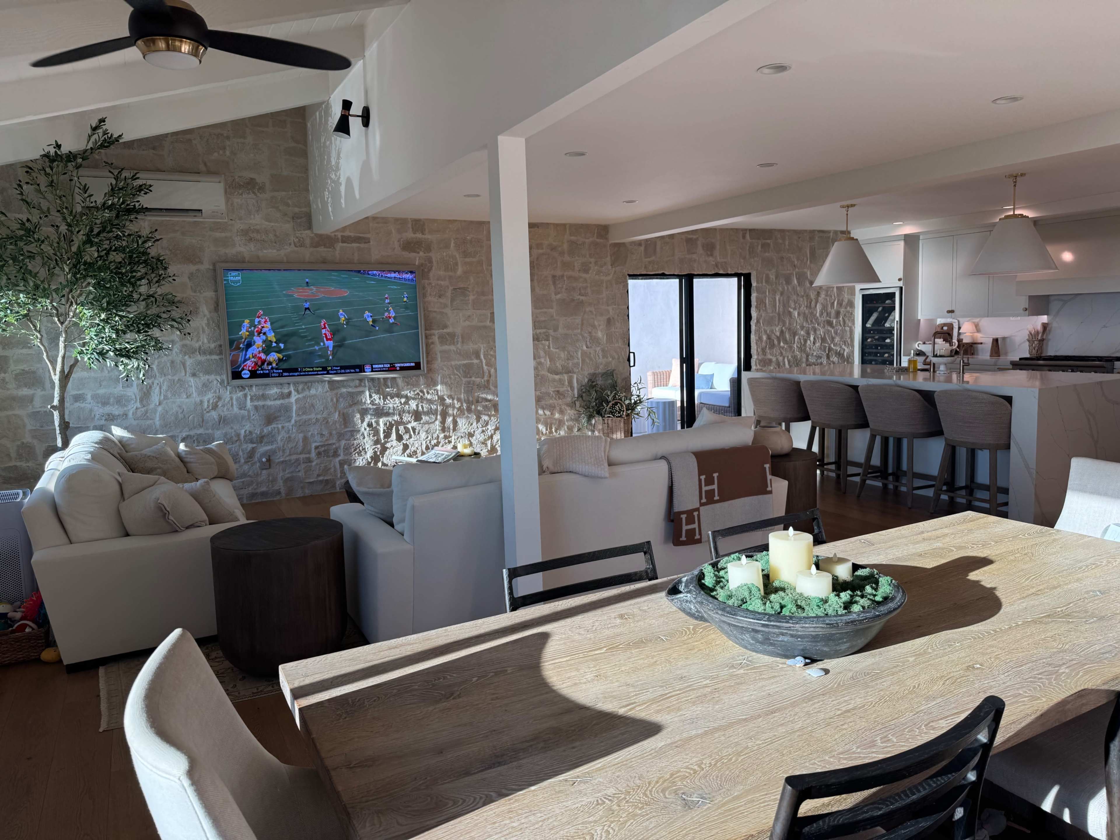 The image shows a modern open-concept living space featuring a dining table, a comfortable seating area, and a kitchen, with a television mounted on the wall displaying a sports game.