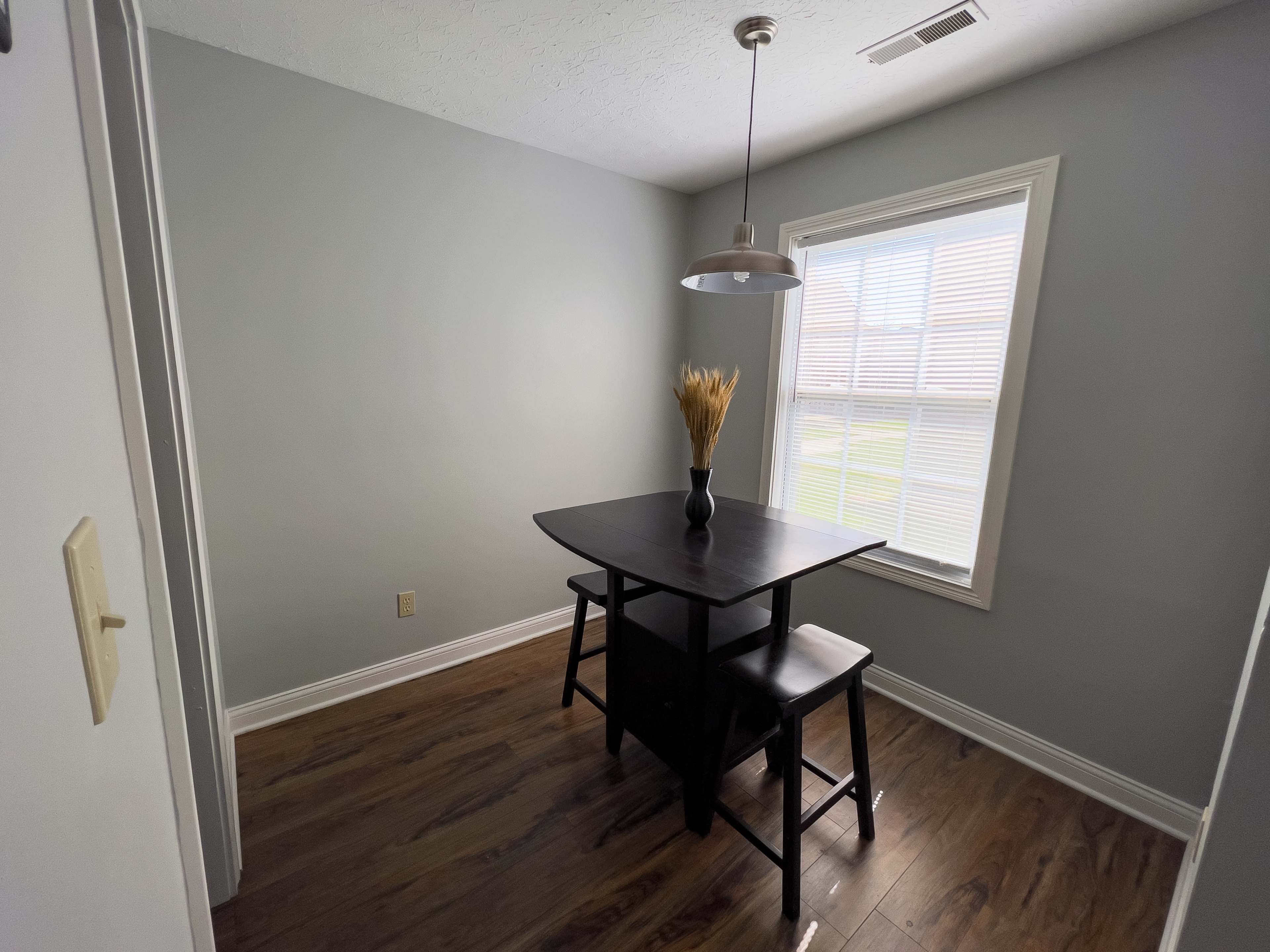 The image shows a small dining area with a black table and a single chair, illuminated by natural light from a nearby window.