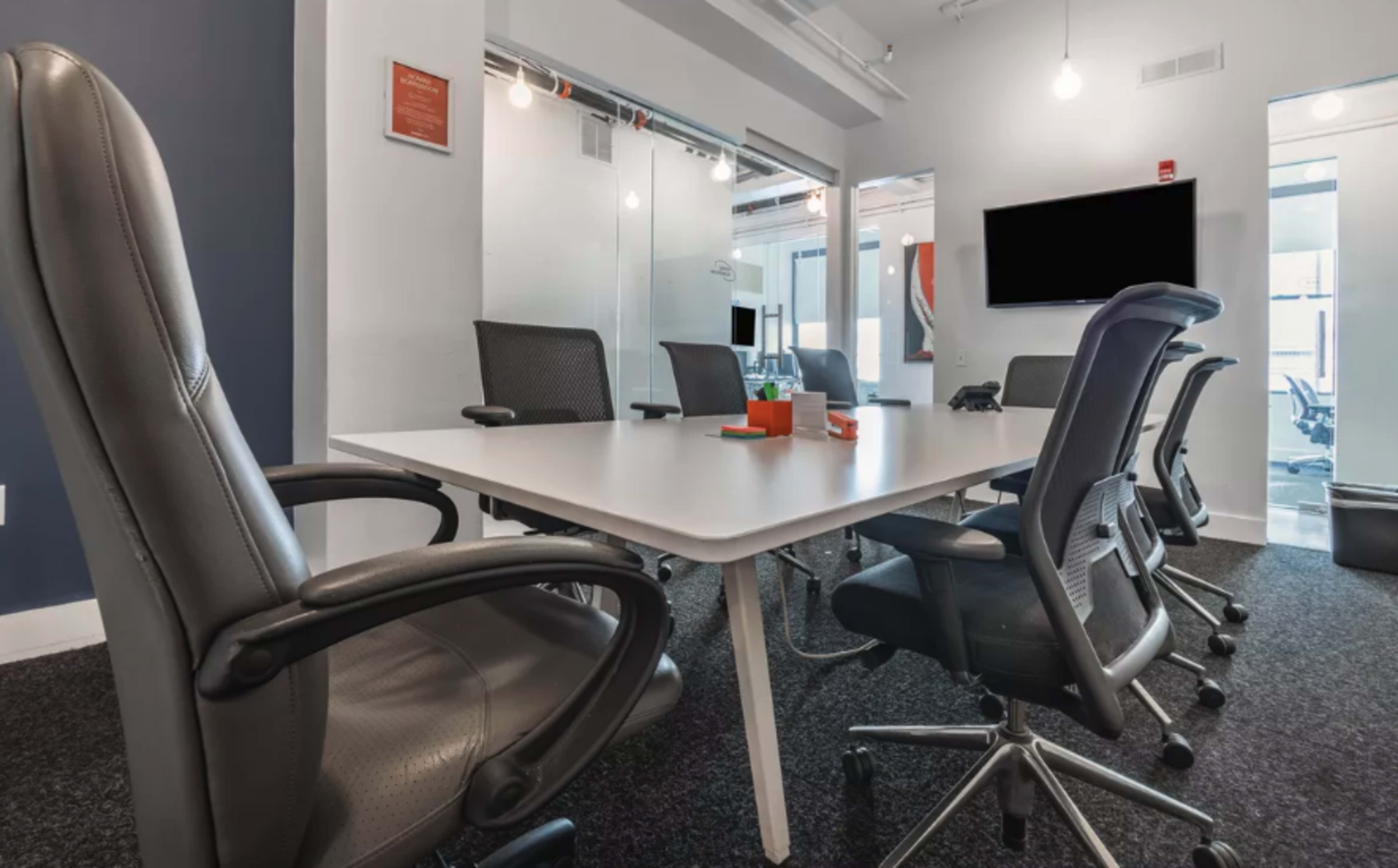 A modern conference room features a large white table surrounded by ergonomic chairs, with a wall-mounted screen and glass-walled meeting space visible.