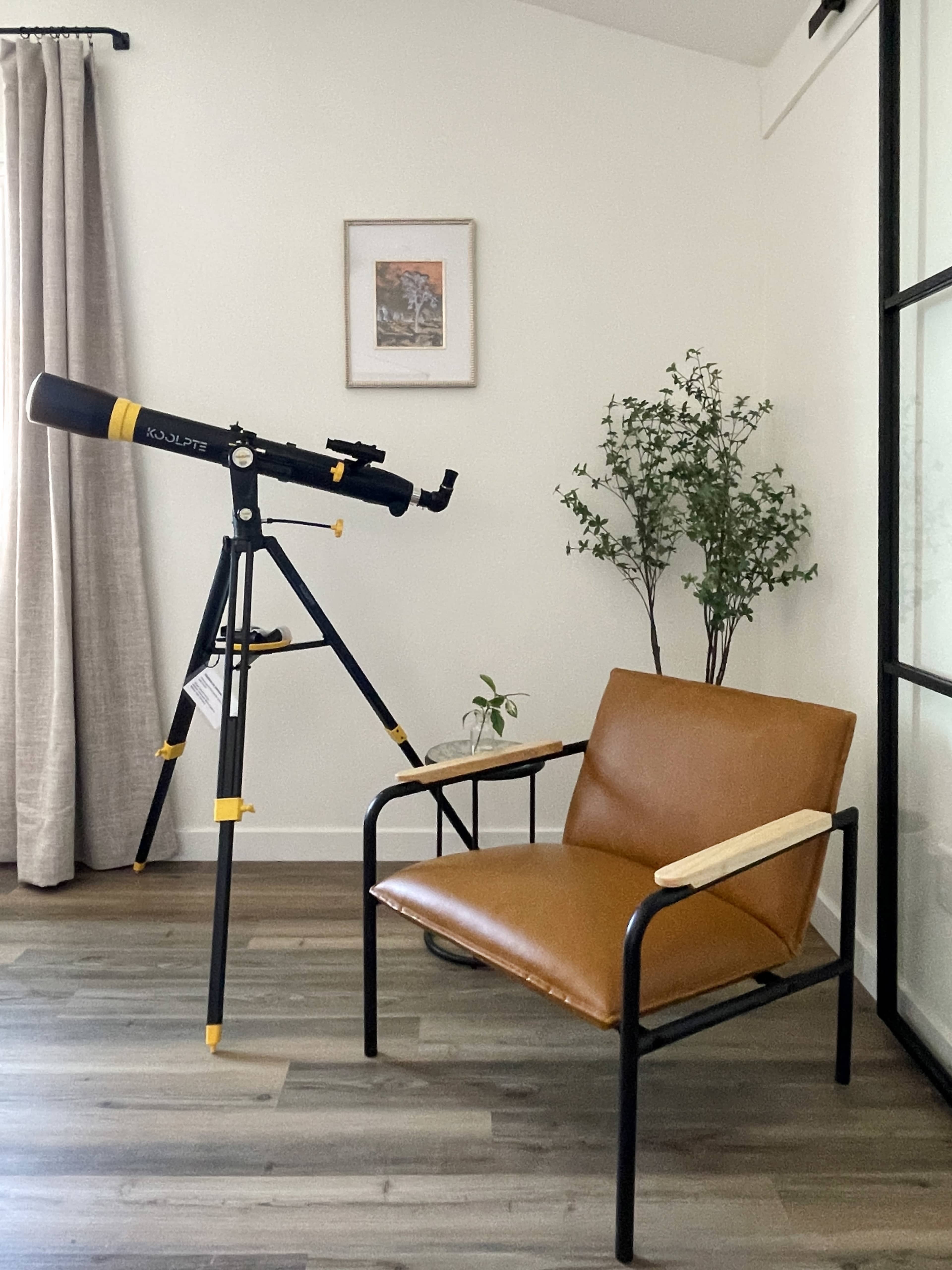 The image shows a telescope positioned next to a brown leather chair in a bright room with a plant and an artwork framed on the wall.