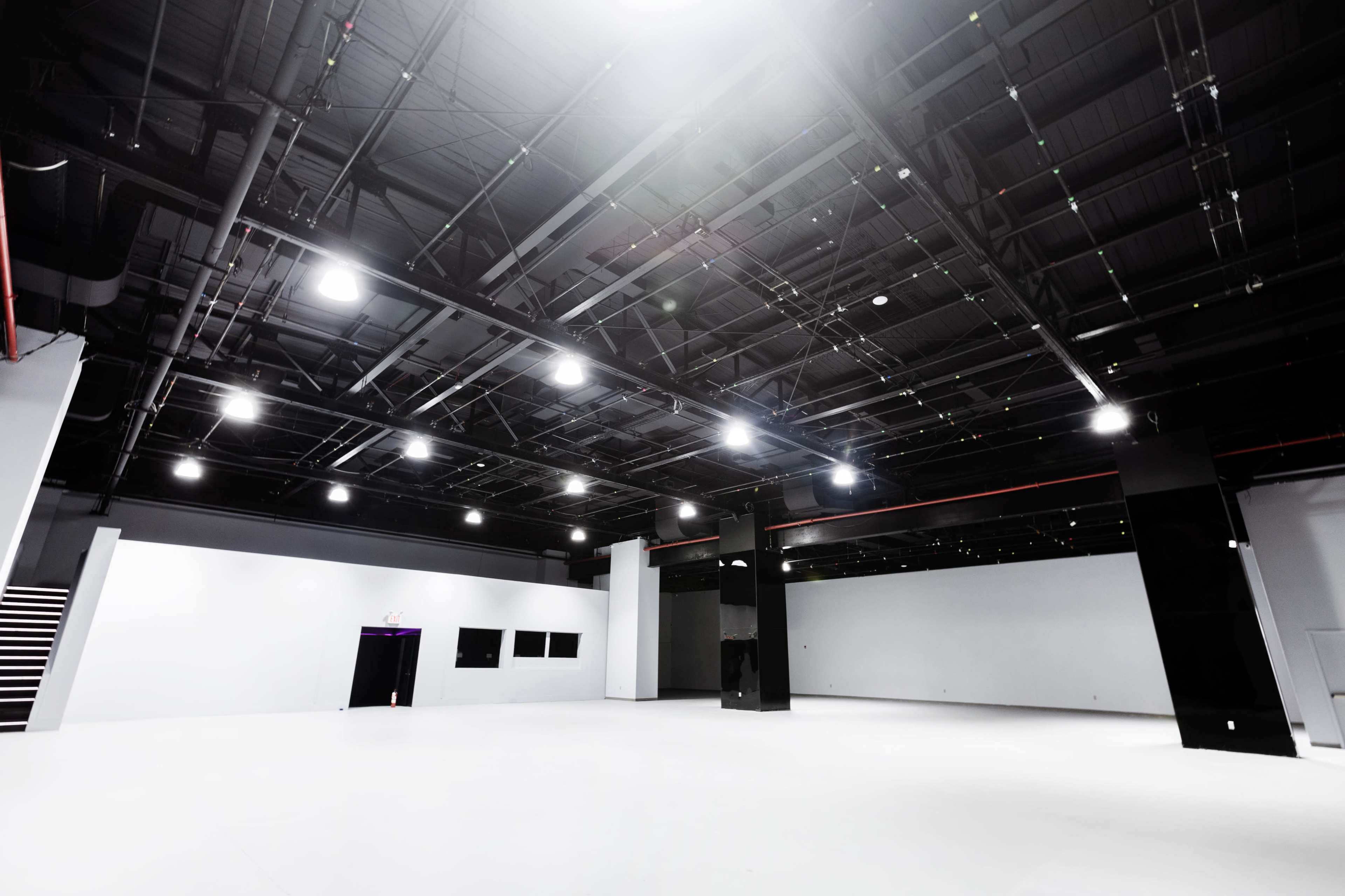 The image shows a spacious, empty interior room with high ceilings, bright overhead lights, and minimal furnishings.