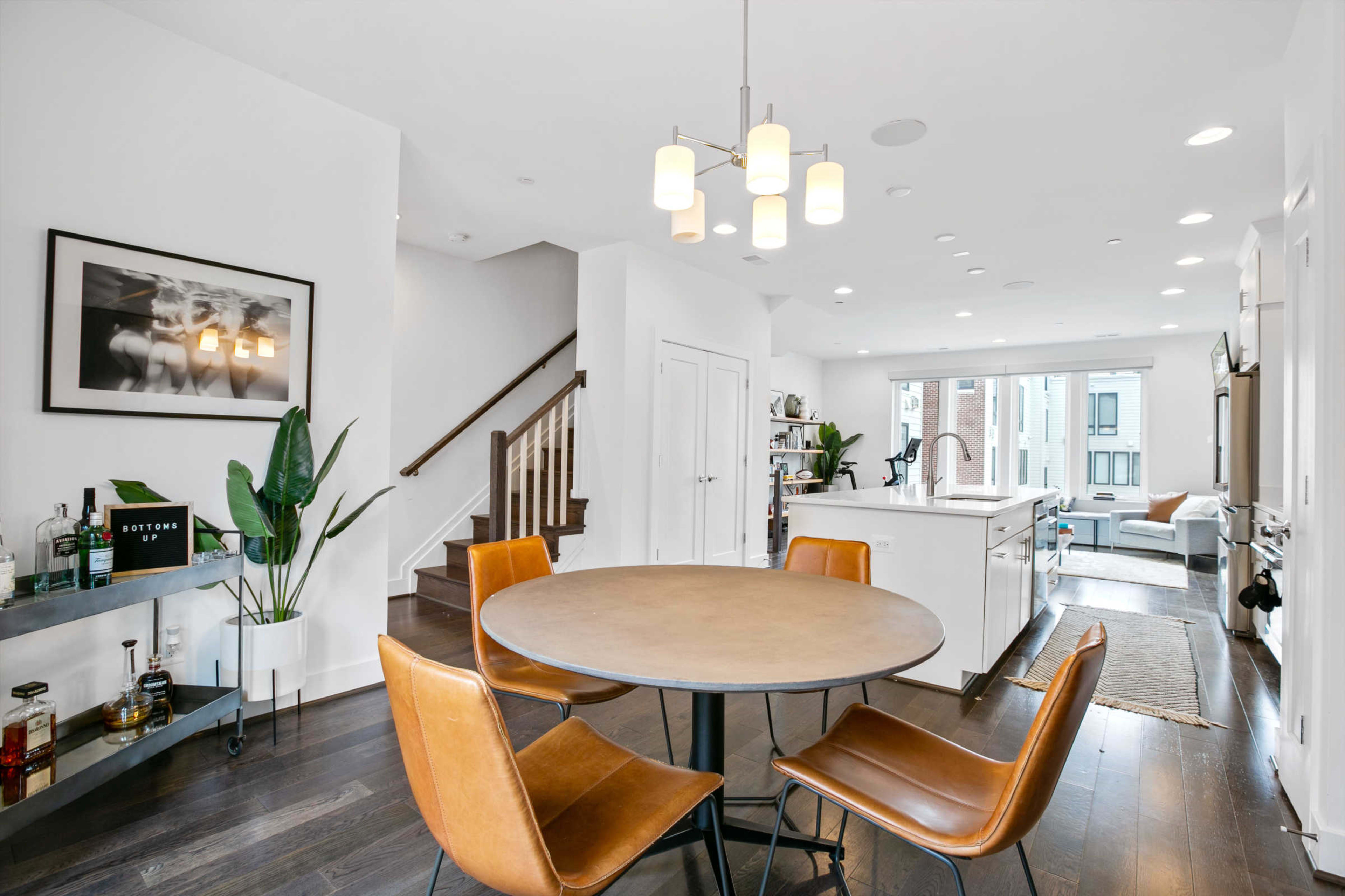 A modern dining area features a round table with brown chairs, adjacent to a staircase and an open kitchen with white cabinetry.