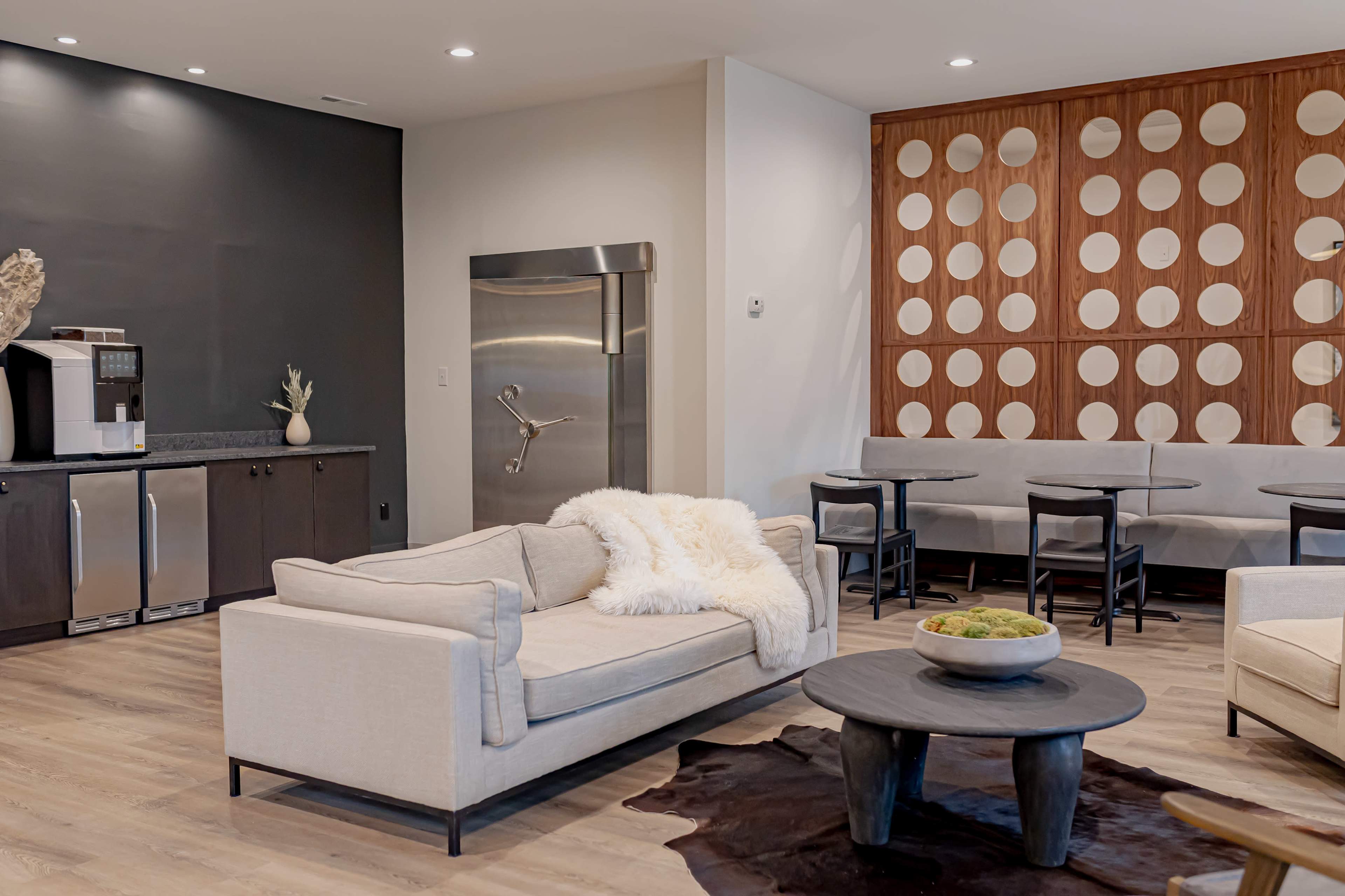 The image shows a modern lounge area featuring a light-colored sofa, a circular wooden table with black chairs, and a kitchenette with a stainless steel refrigerator against a dark wall.