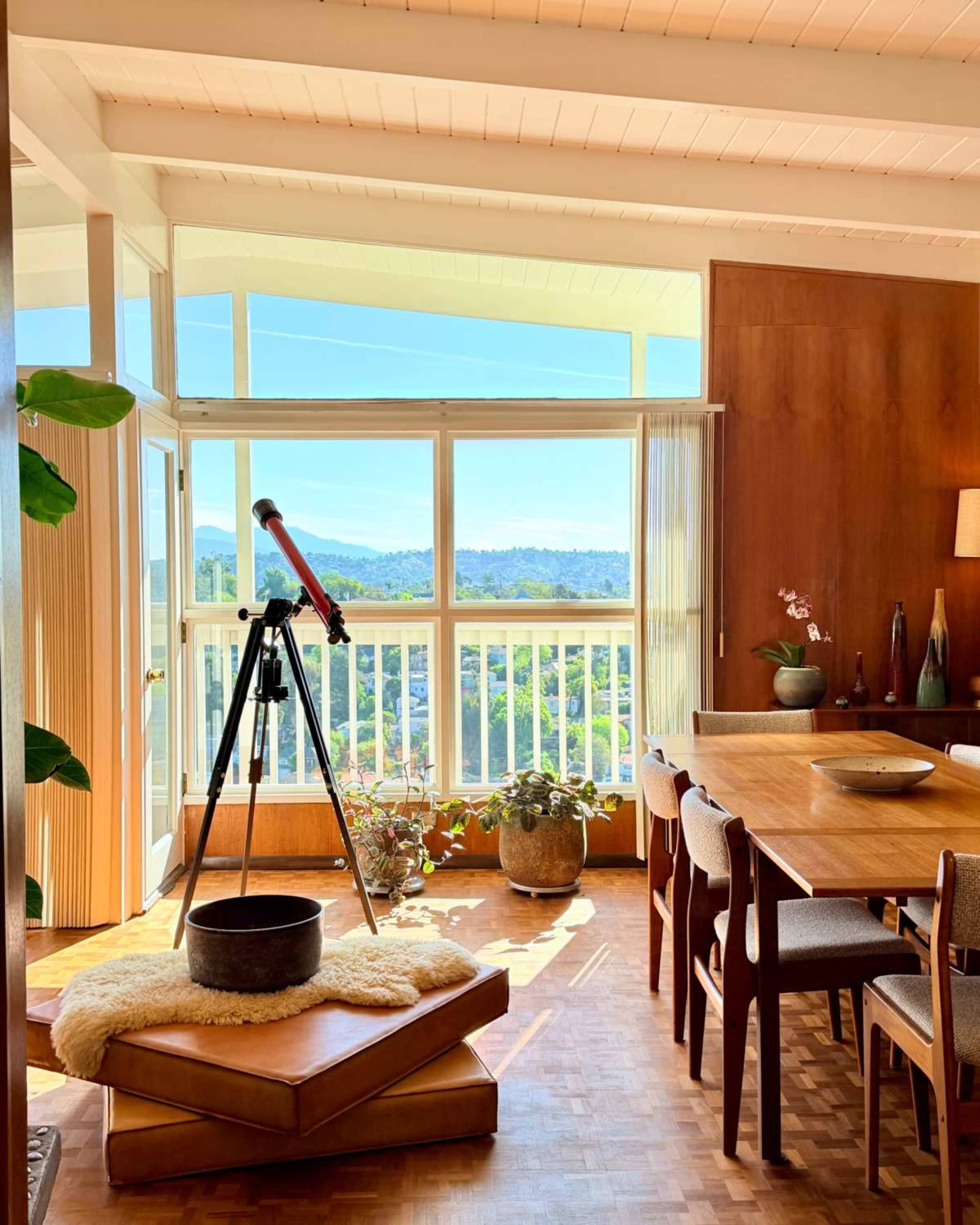 A modern wooden dining area features a telescope beside a large window that provides a view of the landscape outside.