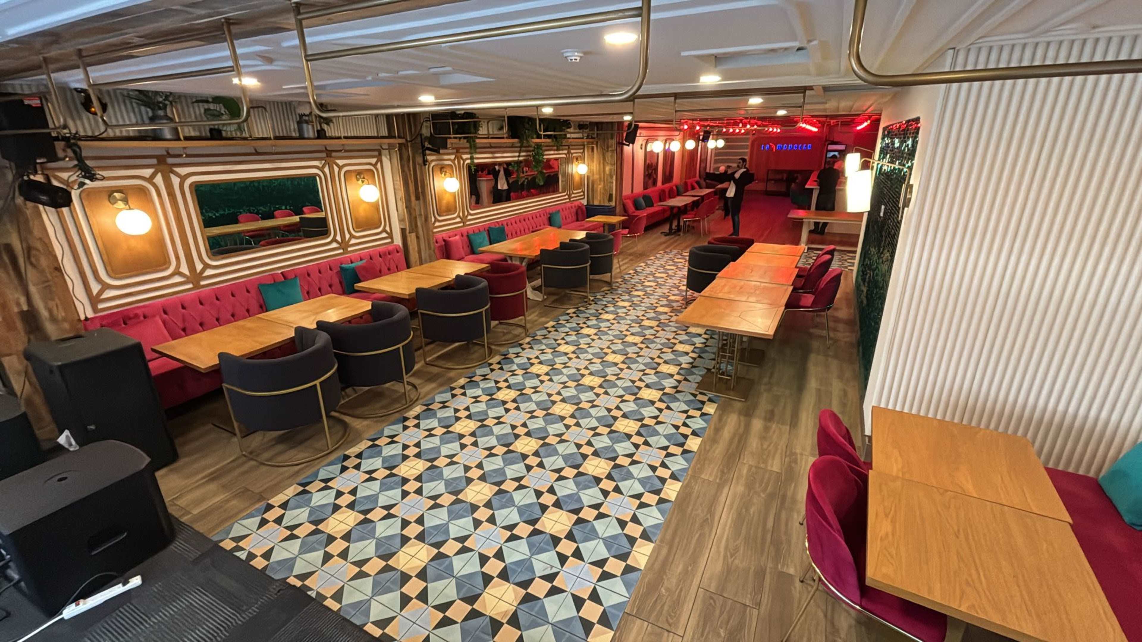 The image shows a spacious restaurant interior with wooden tables, colorful chairs, and patterned flooring, illuminated by warm lighting.