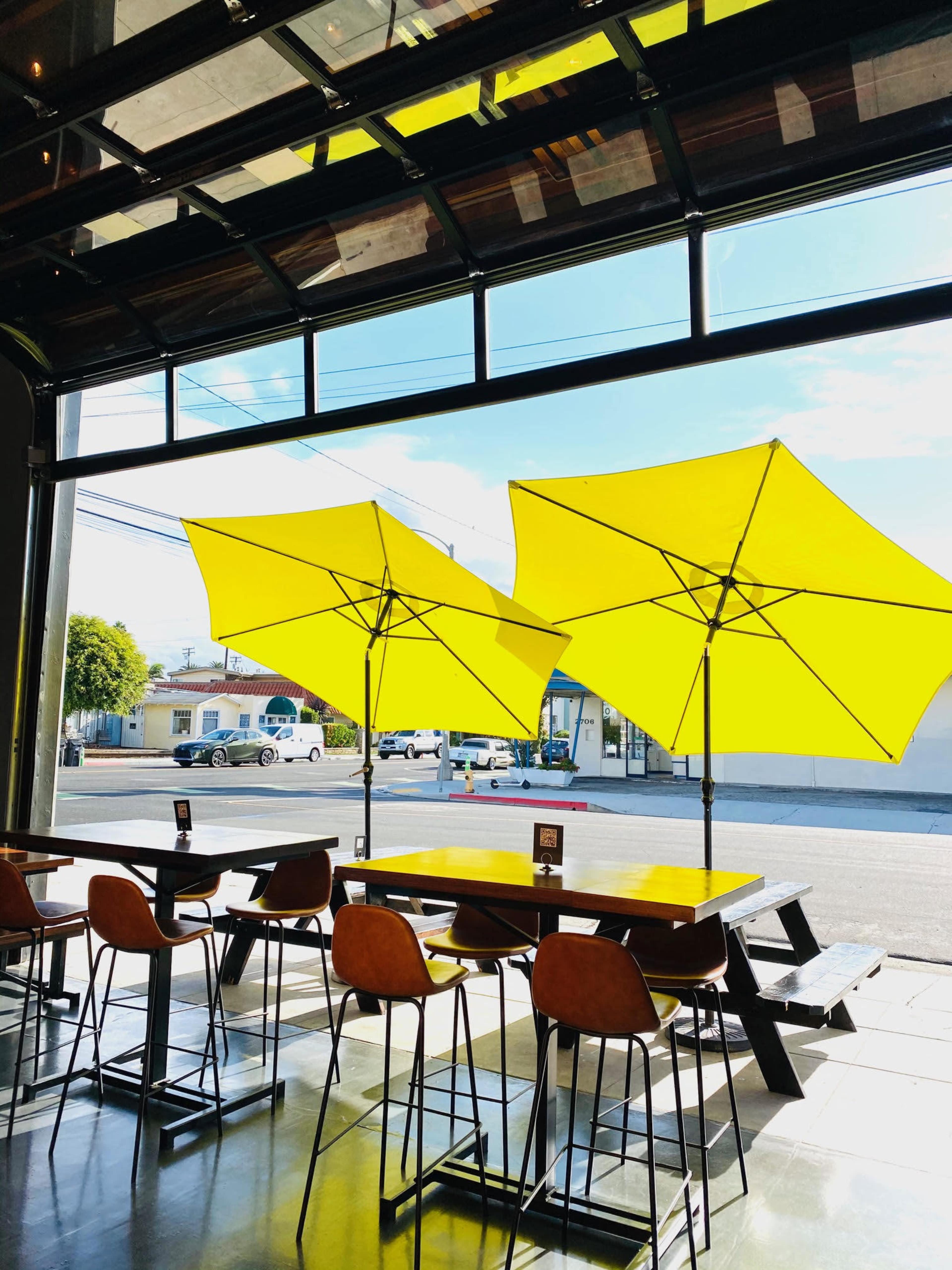 A modern café with yellow umbrellas outside and tall tables arranged inside, illuminated by sunlight.