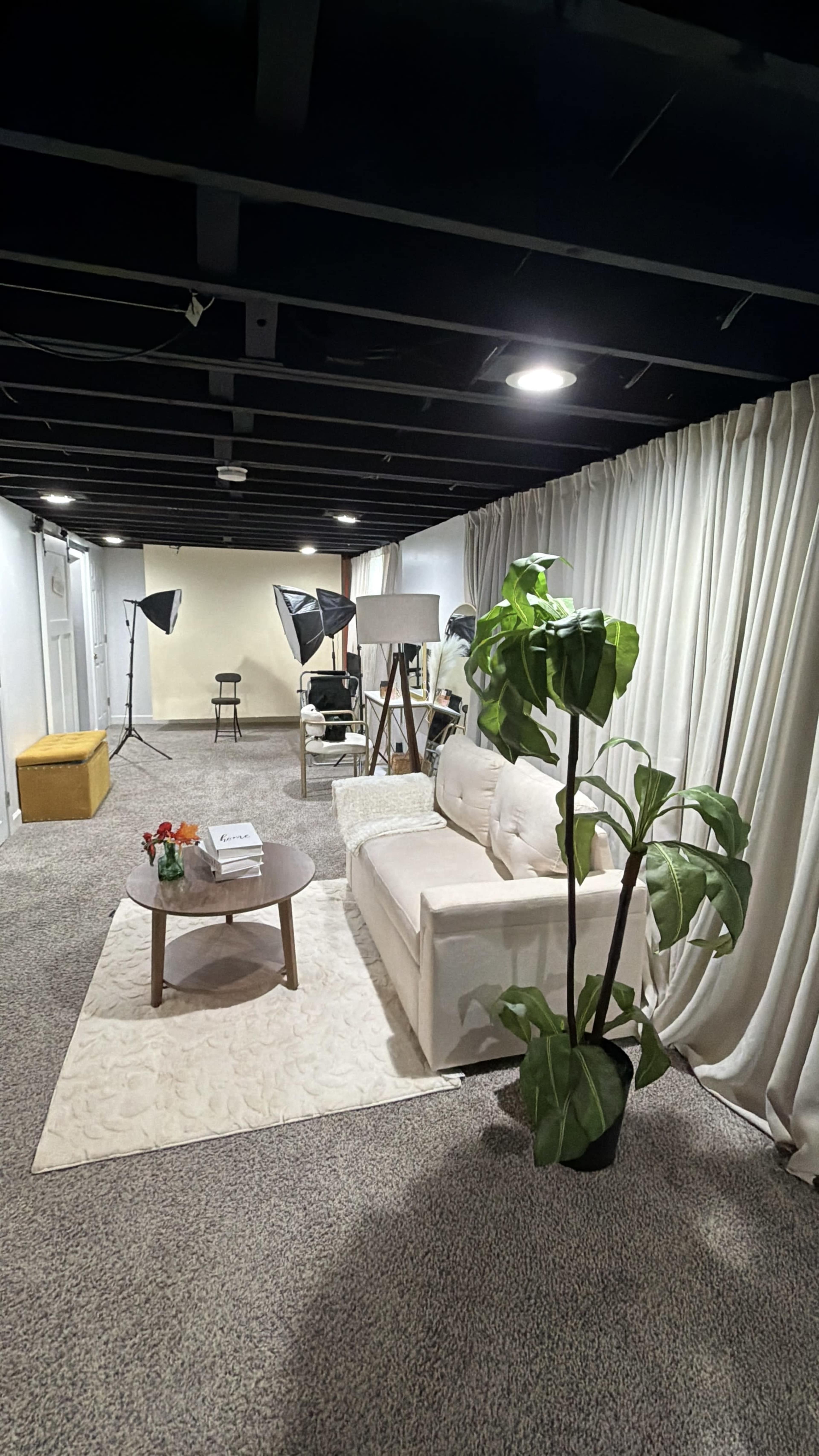 The image shows a well-lit room featuring a white sofa, a coffee table with books, a potted plant, and photography equipment set up against a backdrop of curtains.