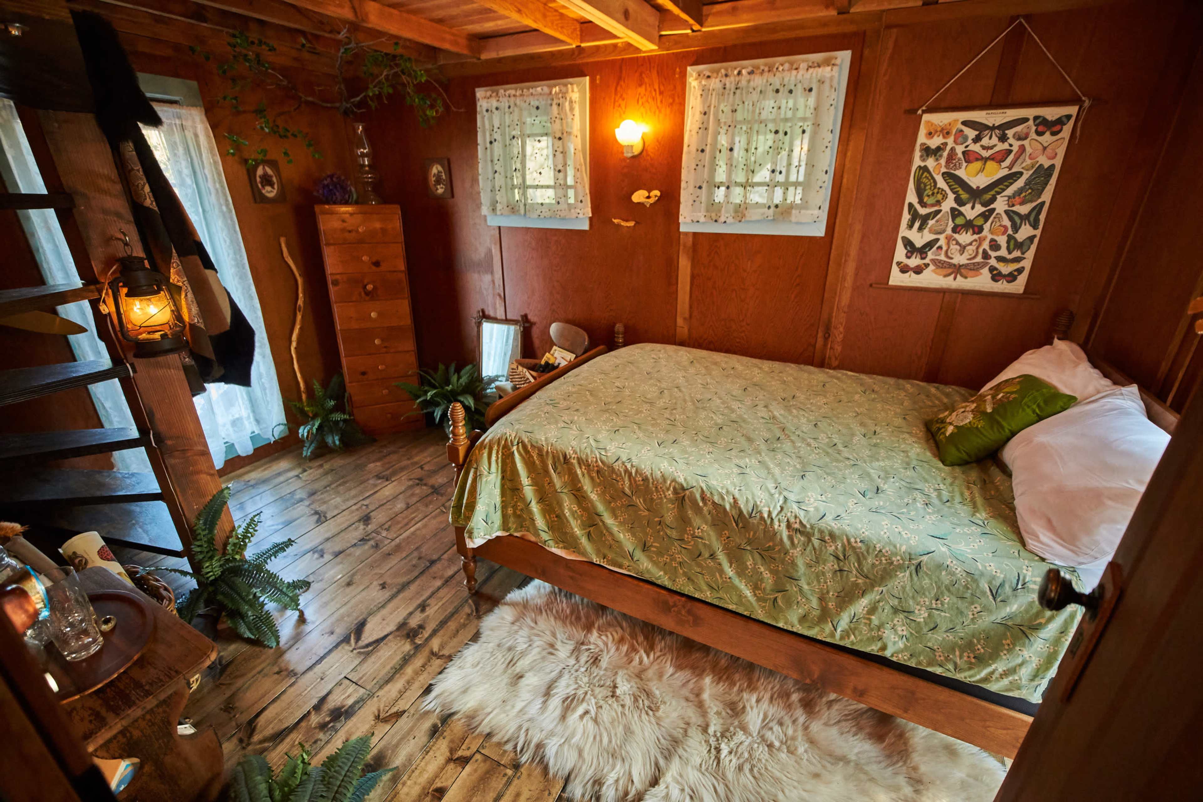 A rustic bedroom with a wooden bed, a furry rug, and various plants, illuminated by warm light from a wall lamp.