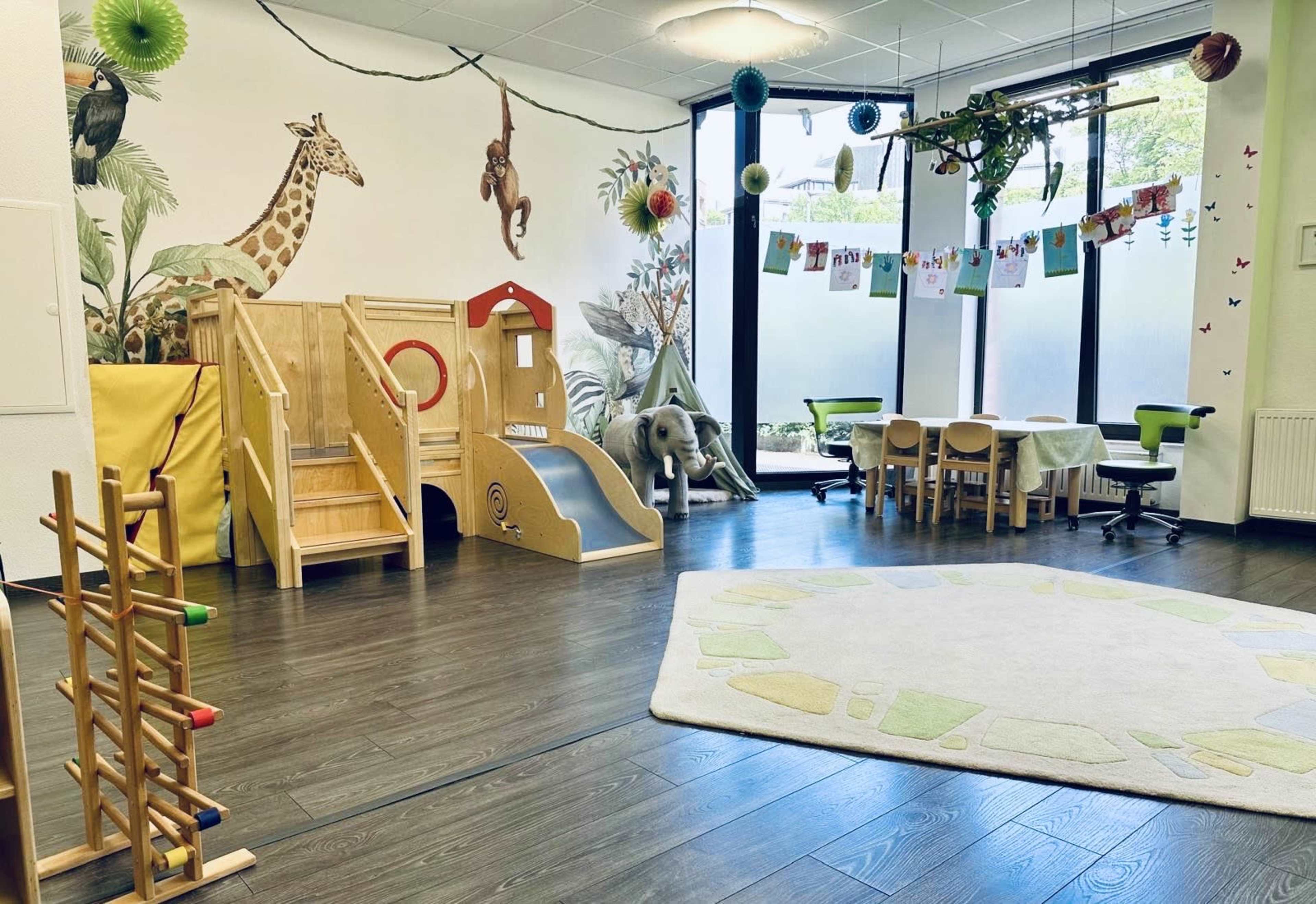 A bright playroom features animal-themed wall murals, wooden play structures, and activity tables arranged around a large area rug.