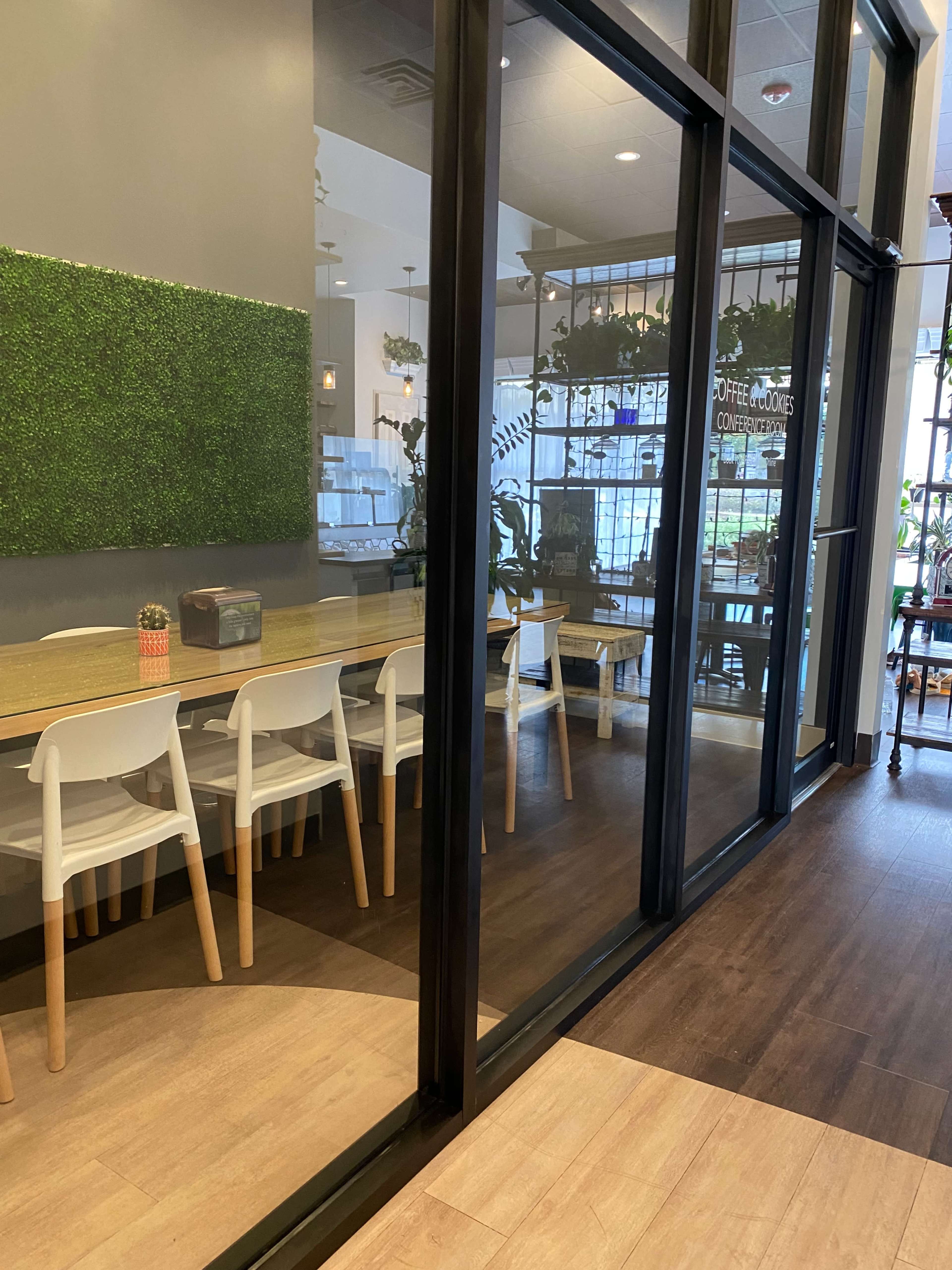 A modern coffee shop with a glass wall, featuring a long wooden table surrounded by white chairs and greenery on the walls.