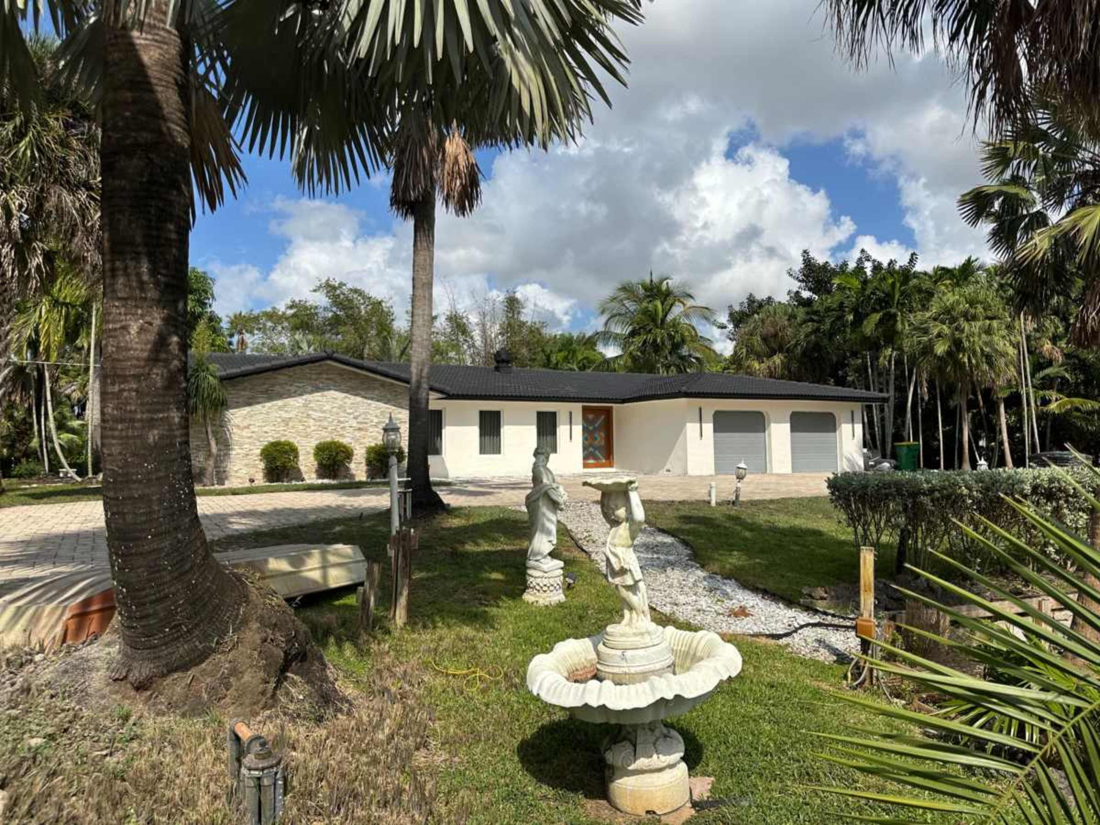 A single-story house with a stone façade and a front porch is surrounded by palm trees and landscaped greenery, featuring a fountain and driveway in the foreground.