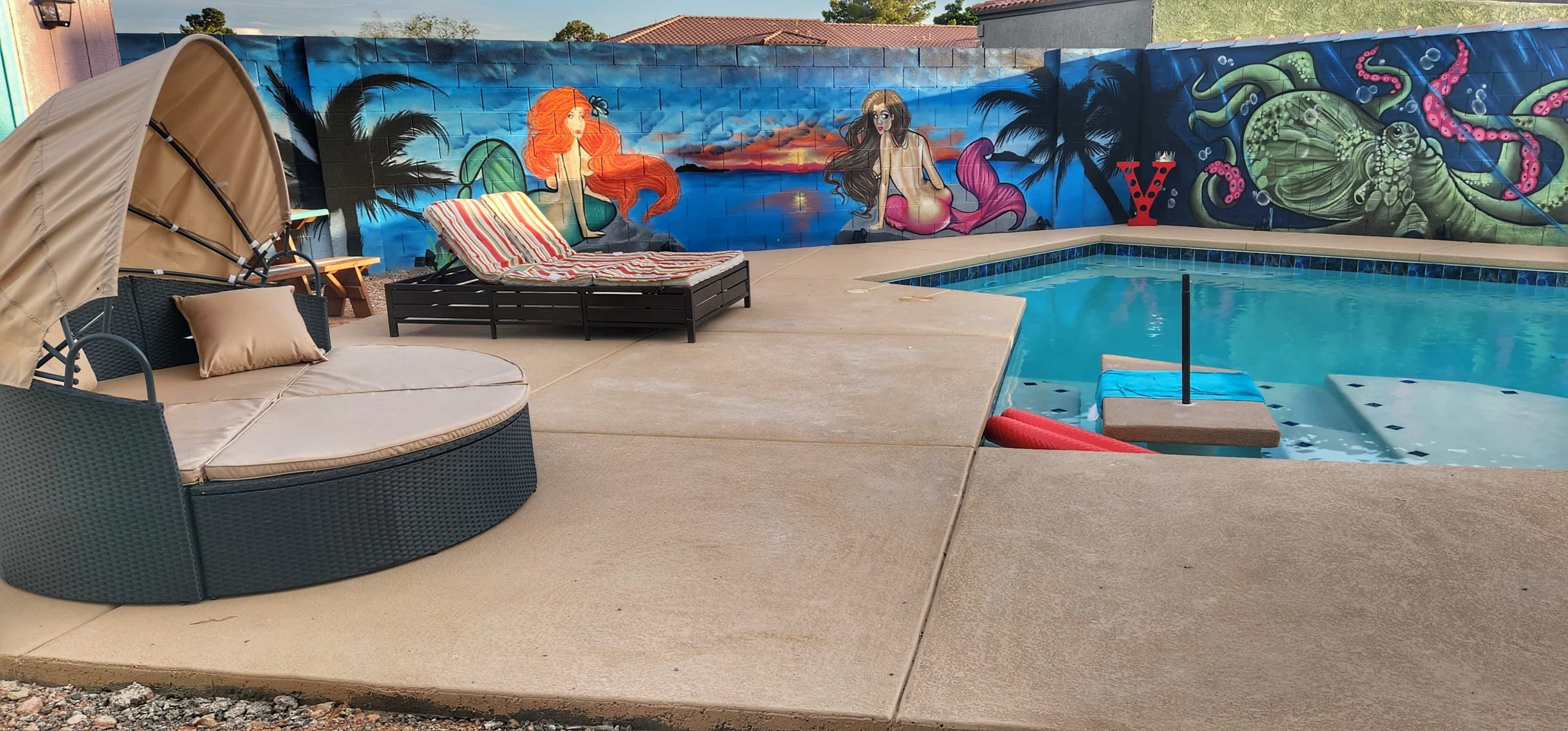 A pool area features a round seating arrangement and lounge chairs beside a colorful mural that includes a mermaid and sea creatures.