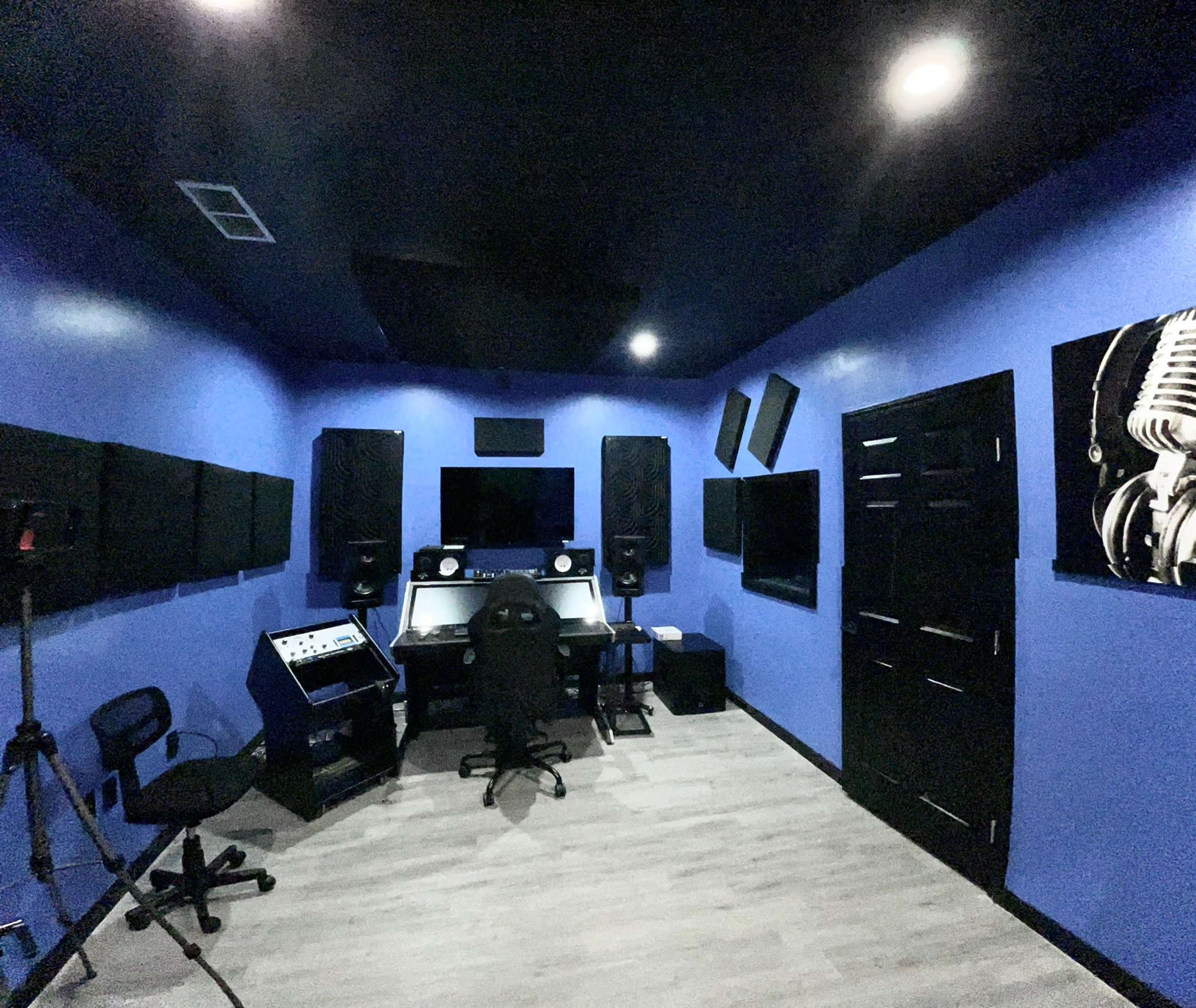 The image shows a recording studio with blue walls, soundproofing panels, and audio equipment set up around a central workstation.
