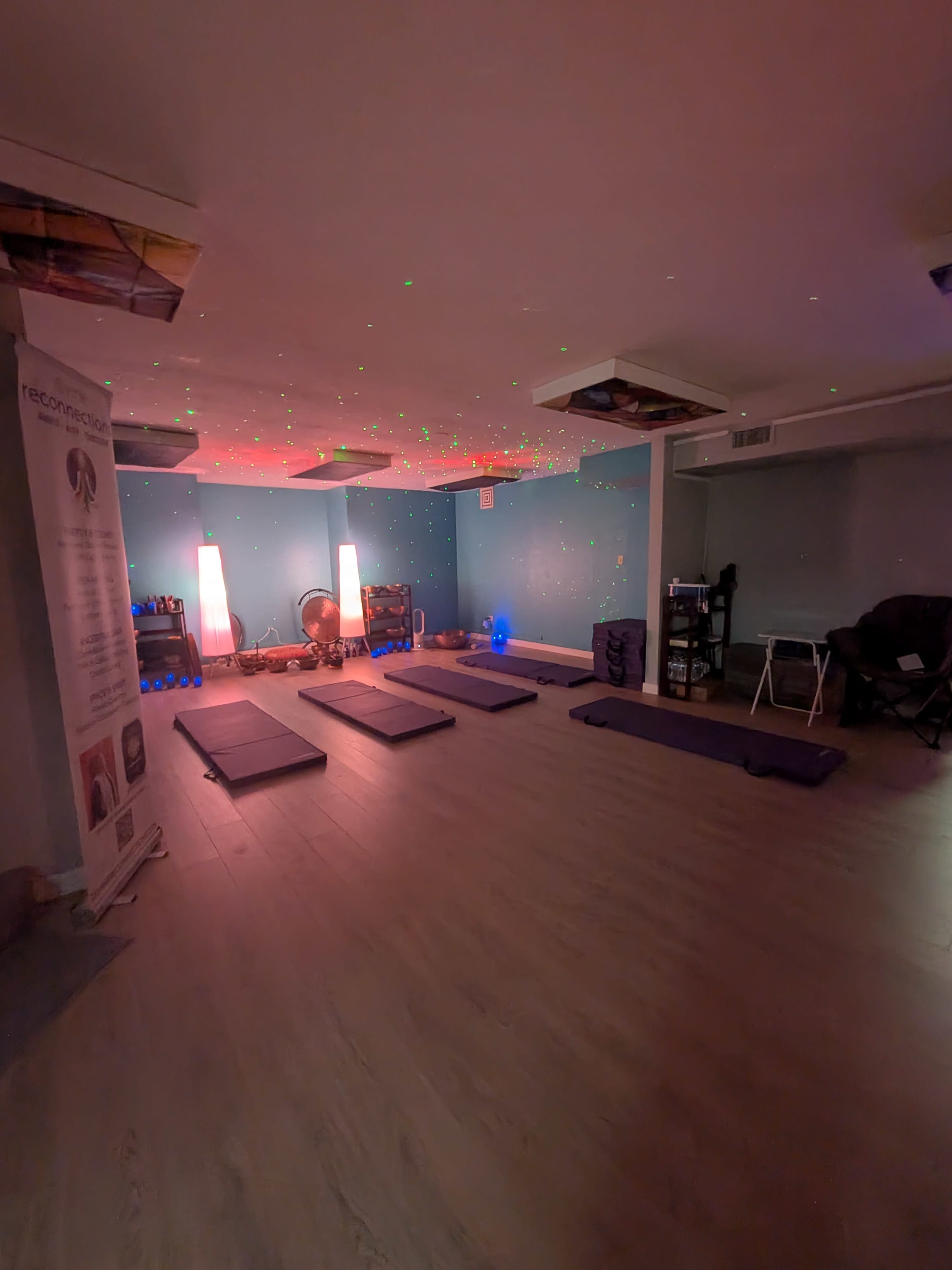 The image shows a softly lit room with yoga mats arranged on a wooden floor, illuminated by colored lights and decorated with calming elements.