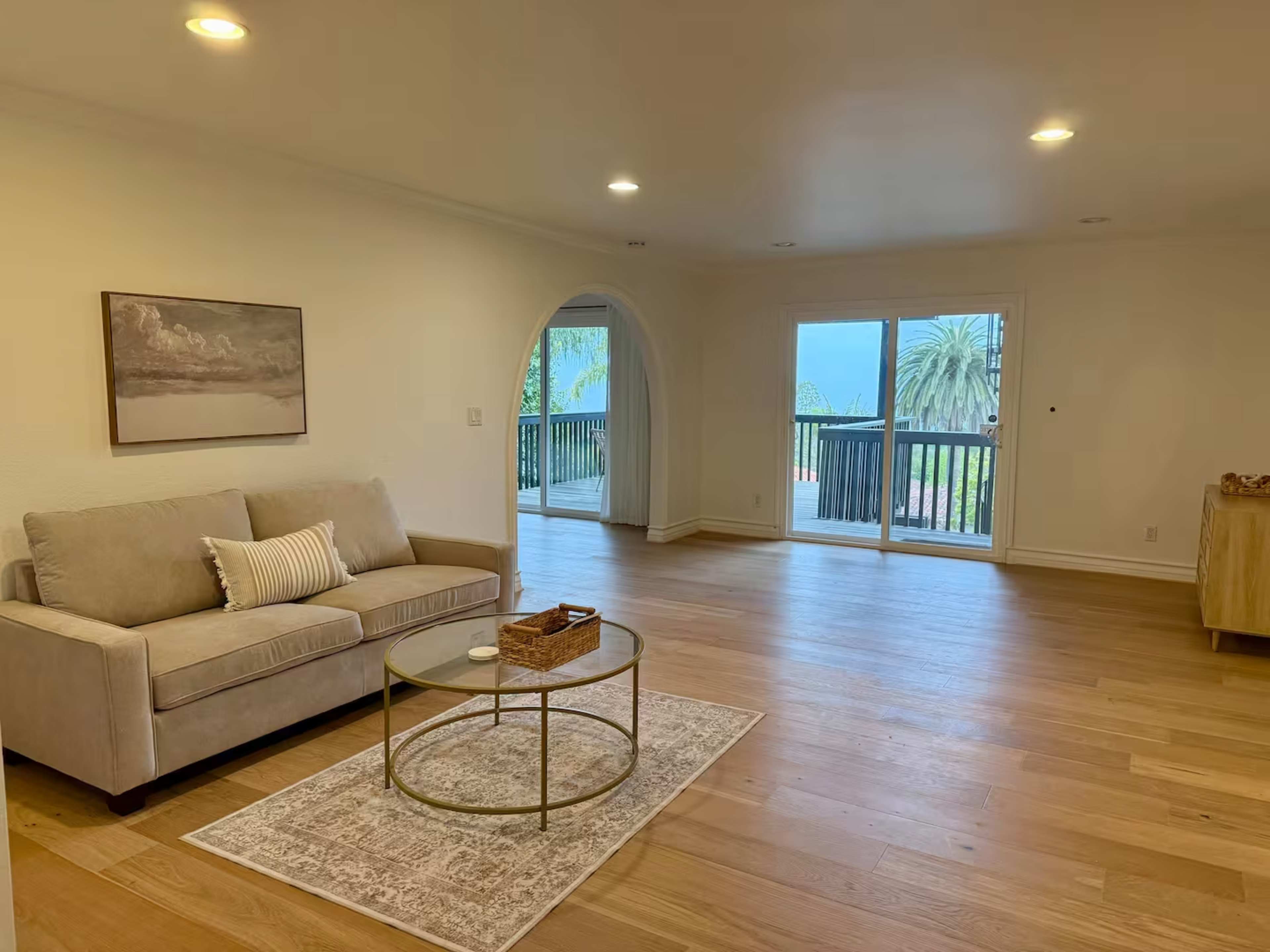 The image shows a spacious living room with a beige sofa, a round coffee table, and large windows that open to a view of trees and a balcony.