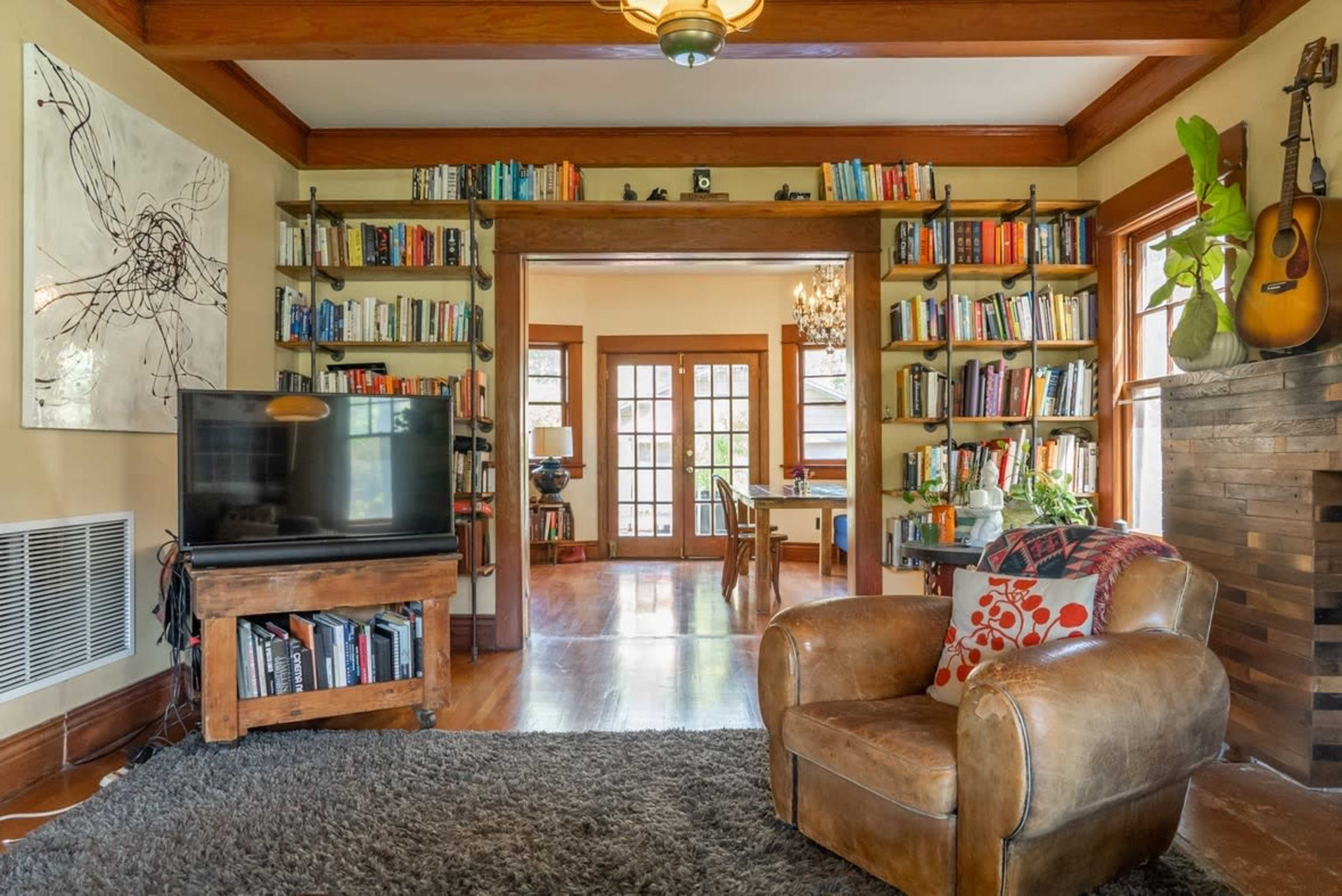 The image shows a cozy living room with bookshelves full of books, a brown leather armchair, a television on a wooden stand, and a view of a dining area through a doorway.