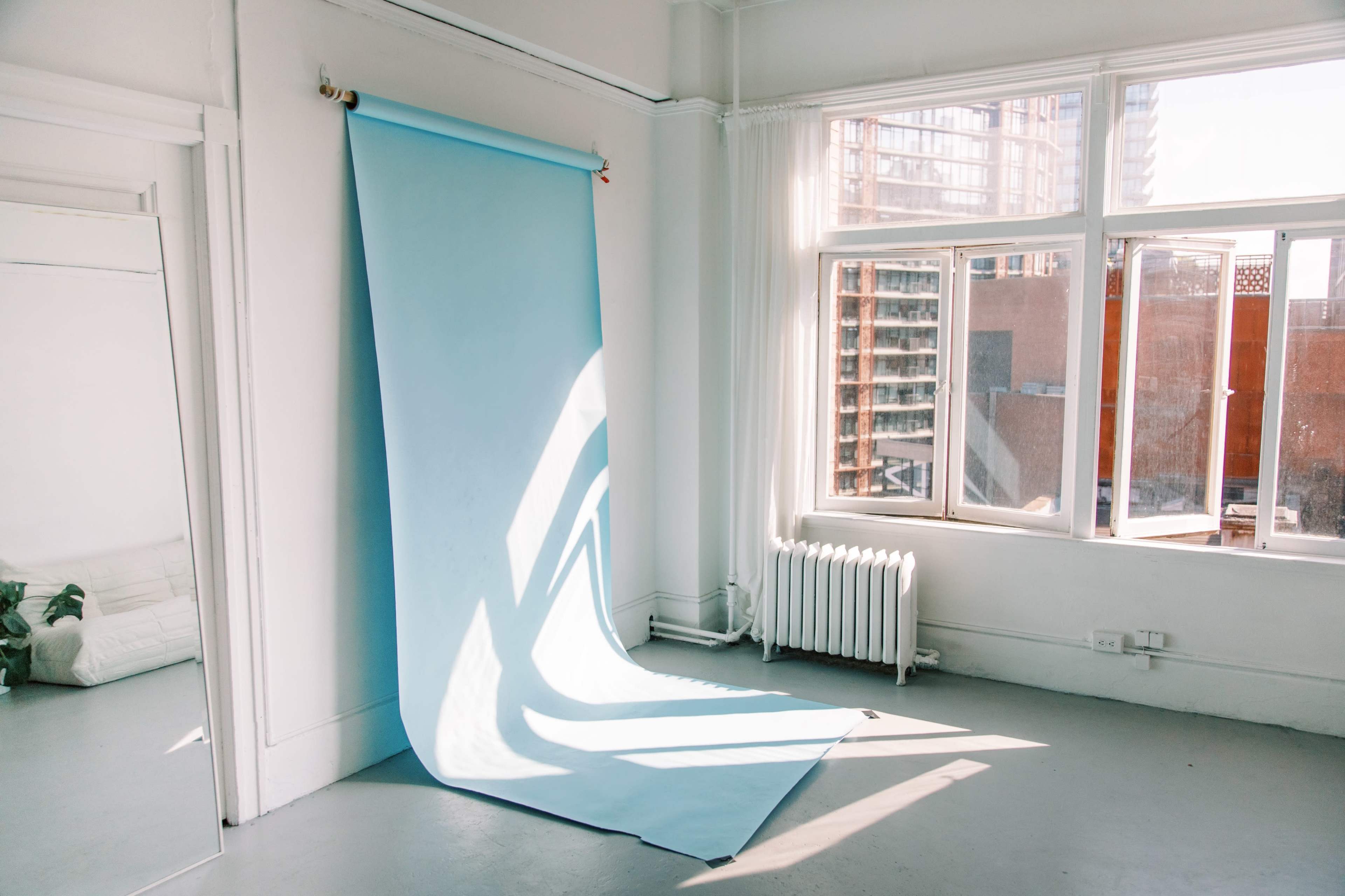 A light blue backdrop against a white wall in a sunlit room with large windows and a mirror.