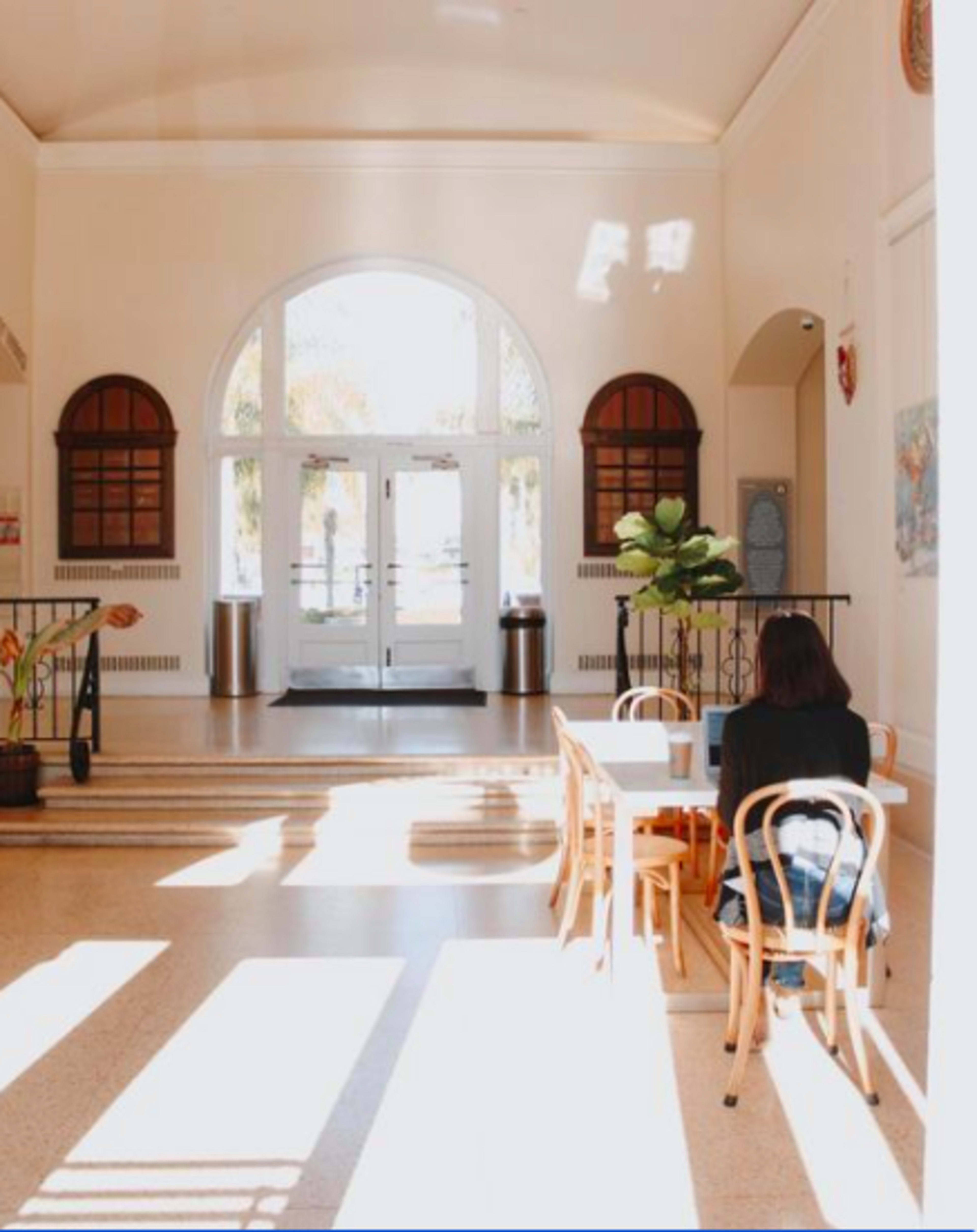 A light-filled entrance hall with a person sitting at a table, facing a set of large doors.