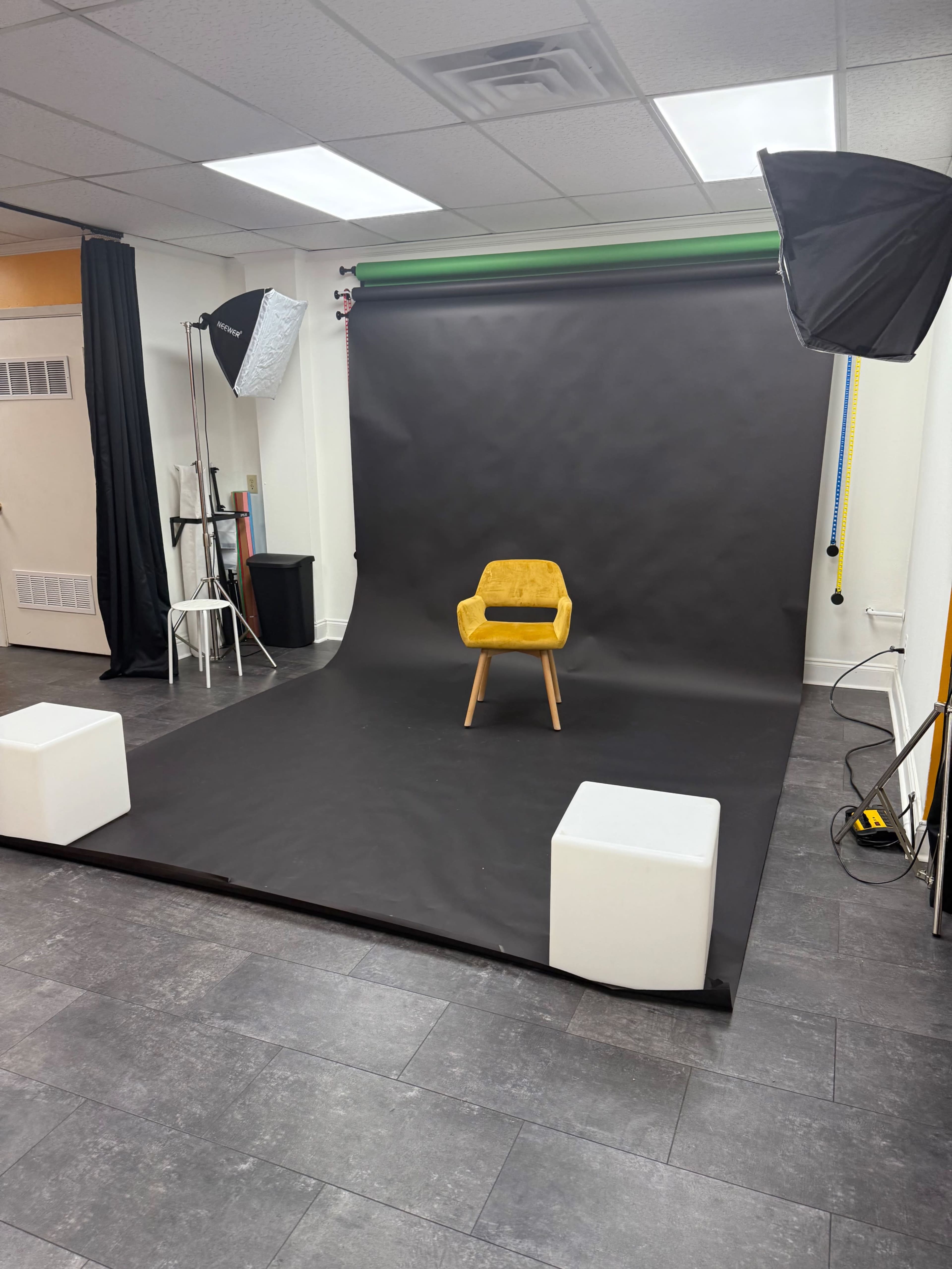 The image shows a photography studio setup with a black backdrop, a yellow chair in the center, and two white cube props on the floor.