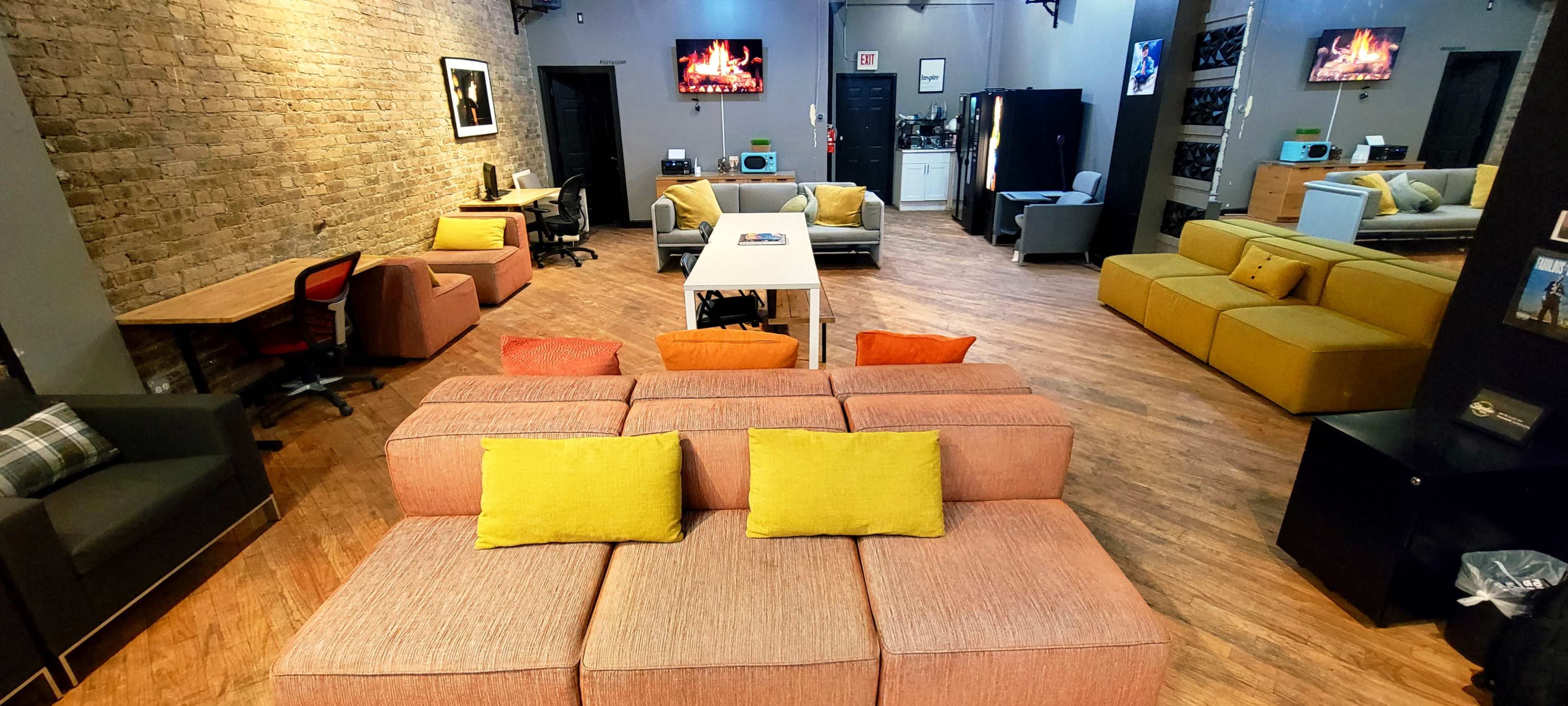 The image shows a modern, spacious lounge area with various seating arrangements, including a modular sofa with bright cushions, desks, and a small kitchenette in the background.