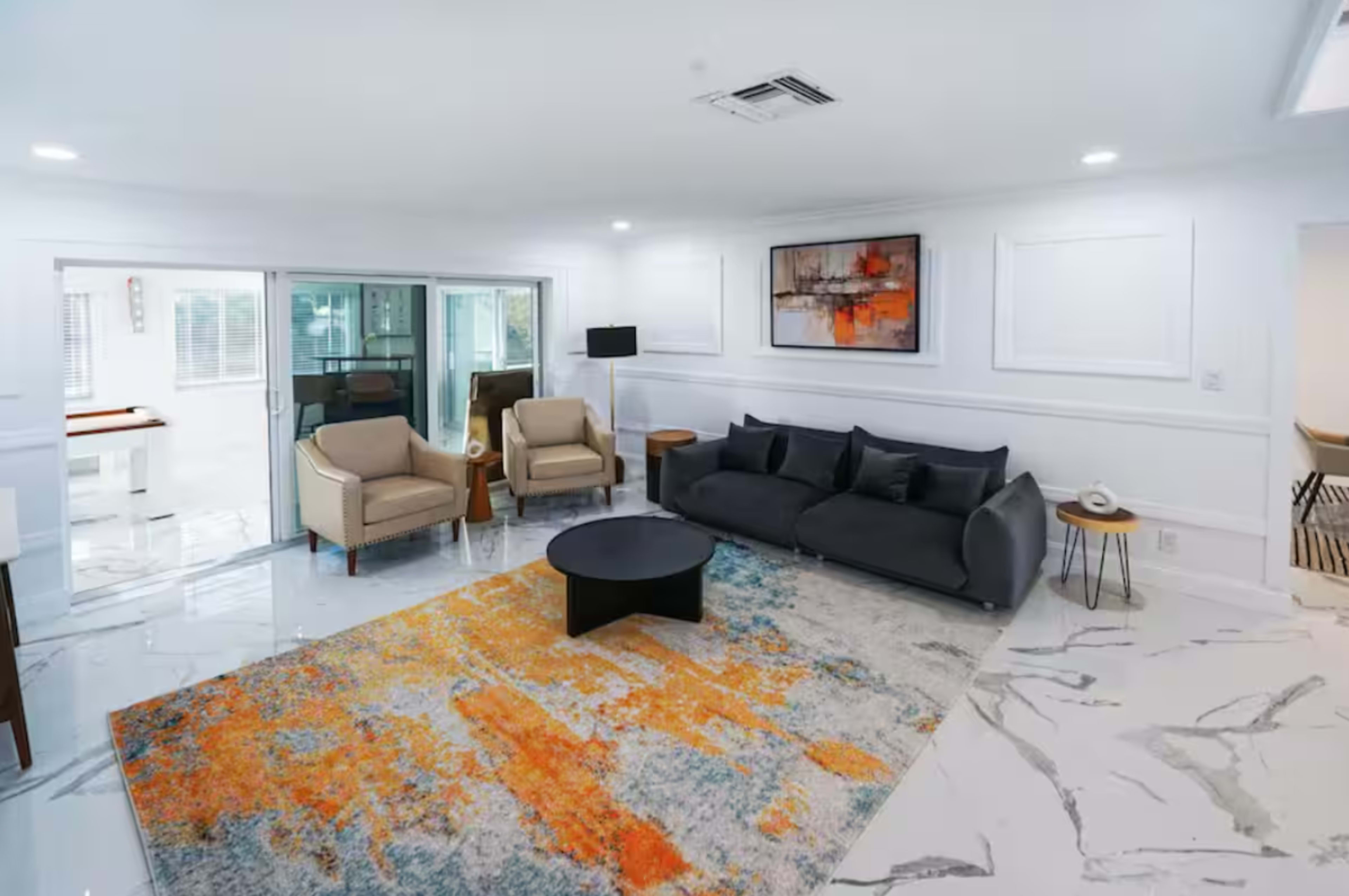 The image shows a modern living room featuring a gray sofa, two beige armchairs, and a round black coffee table on a colorful area rug, with large windows allowing natural light.
