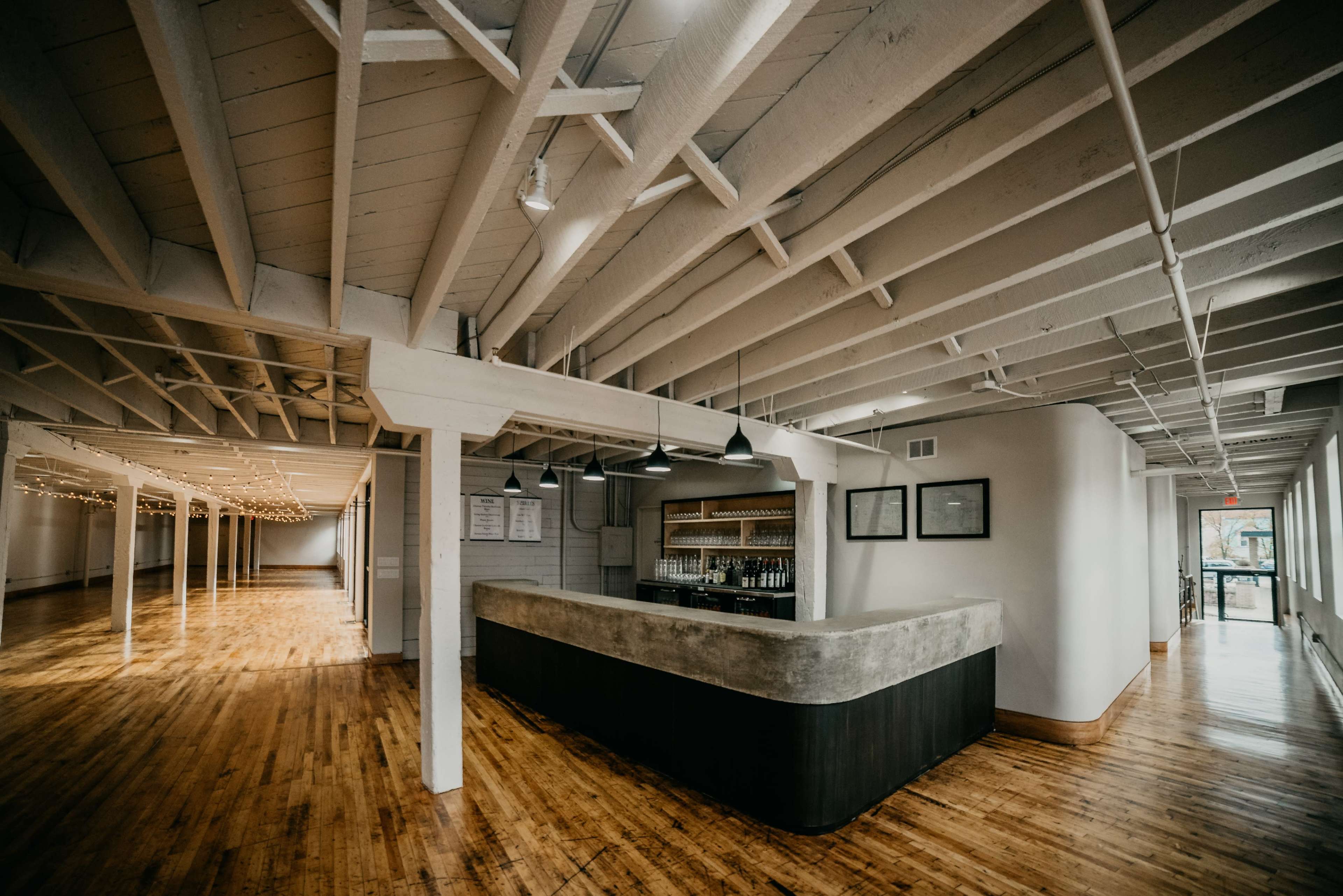 The image shows a spacious, well-lit interior with polished wooden floors and a bar area featuring a concrete counter.