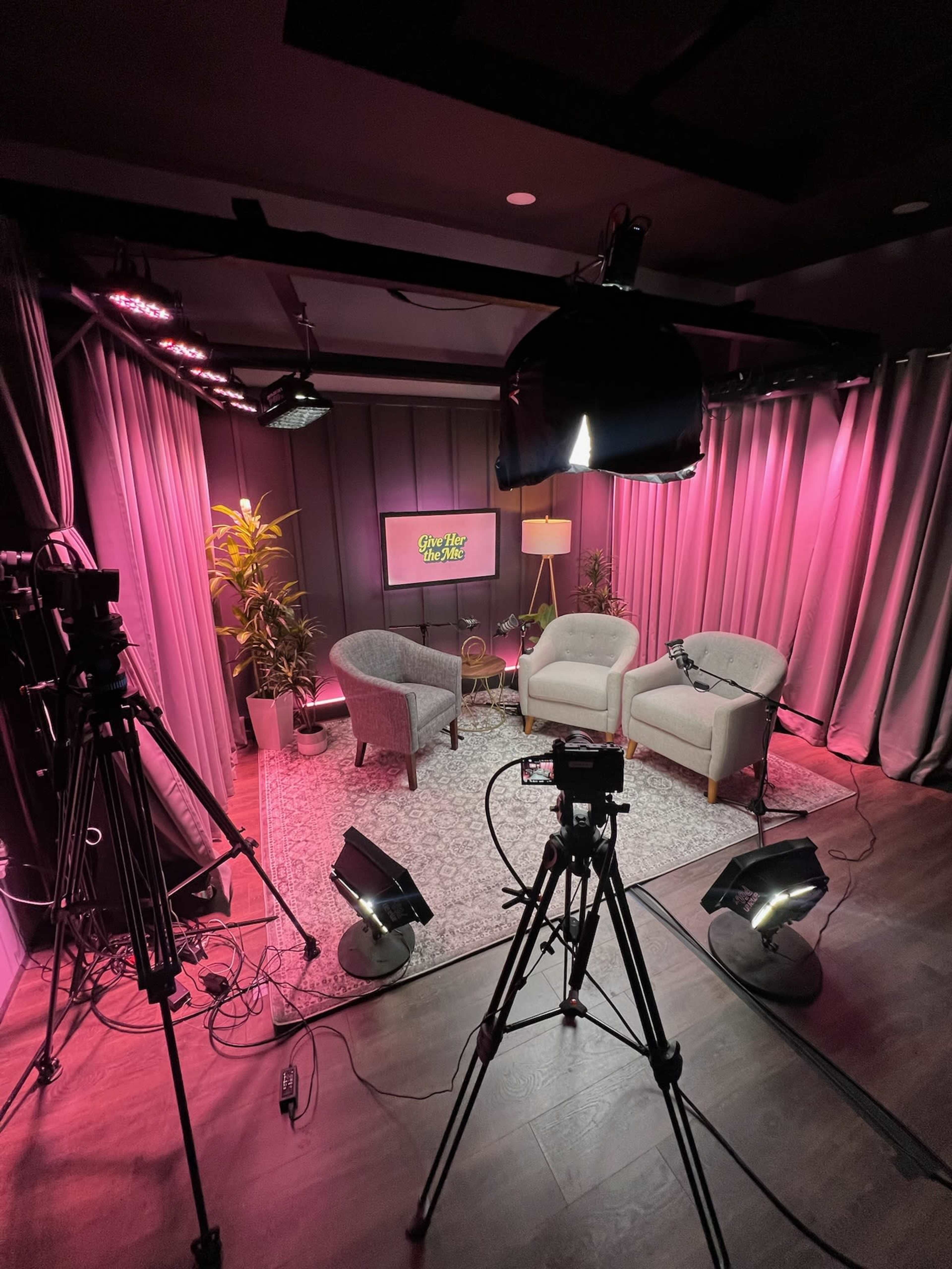 The image shows a brightly lit small studio setup with two chairs, a coffee table, and backdrop curtains, alongside cameras and lights arranged for video recording.