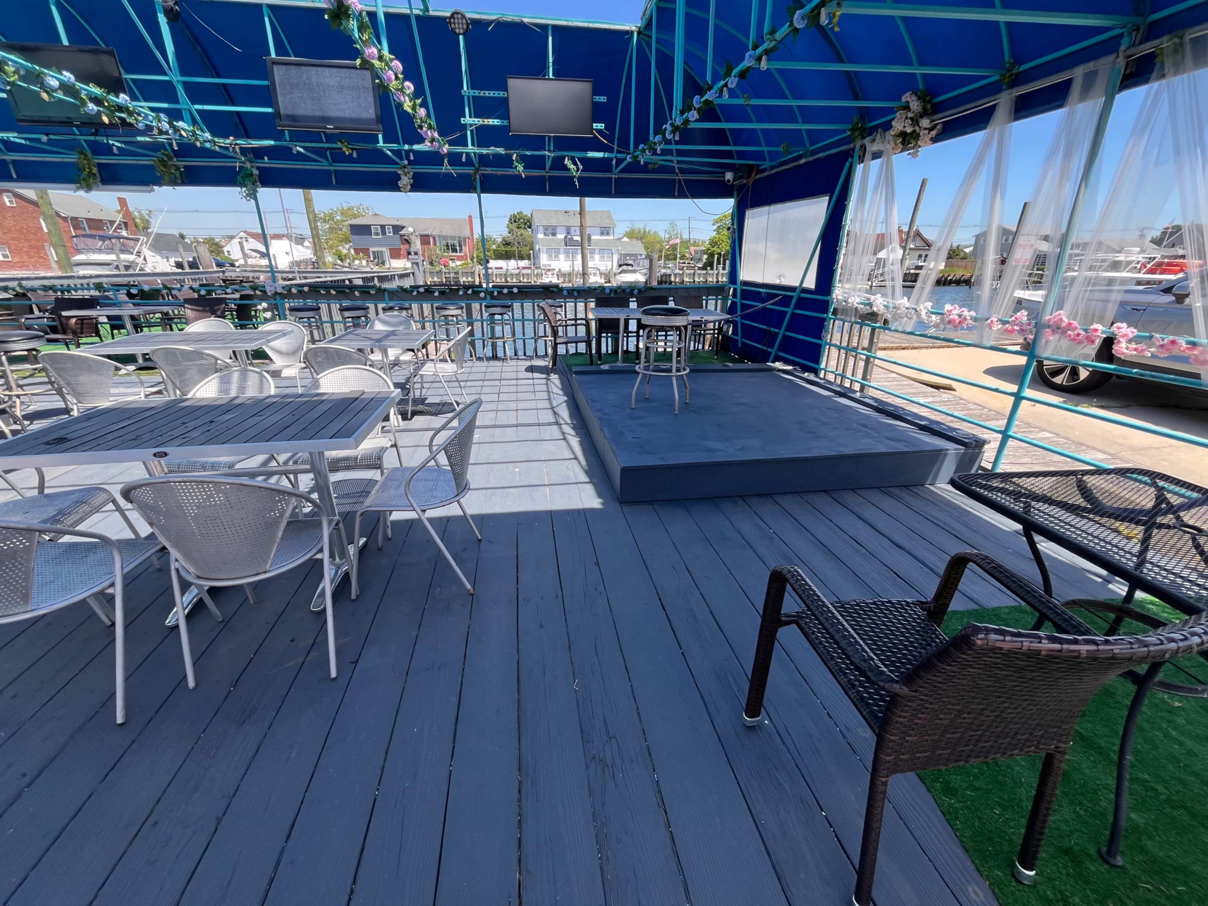 The image shows a large outdoor dining area with several tables and chairs, a small elevated stage, and blue canopies overhead.