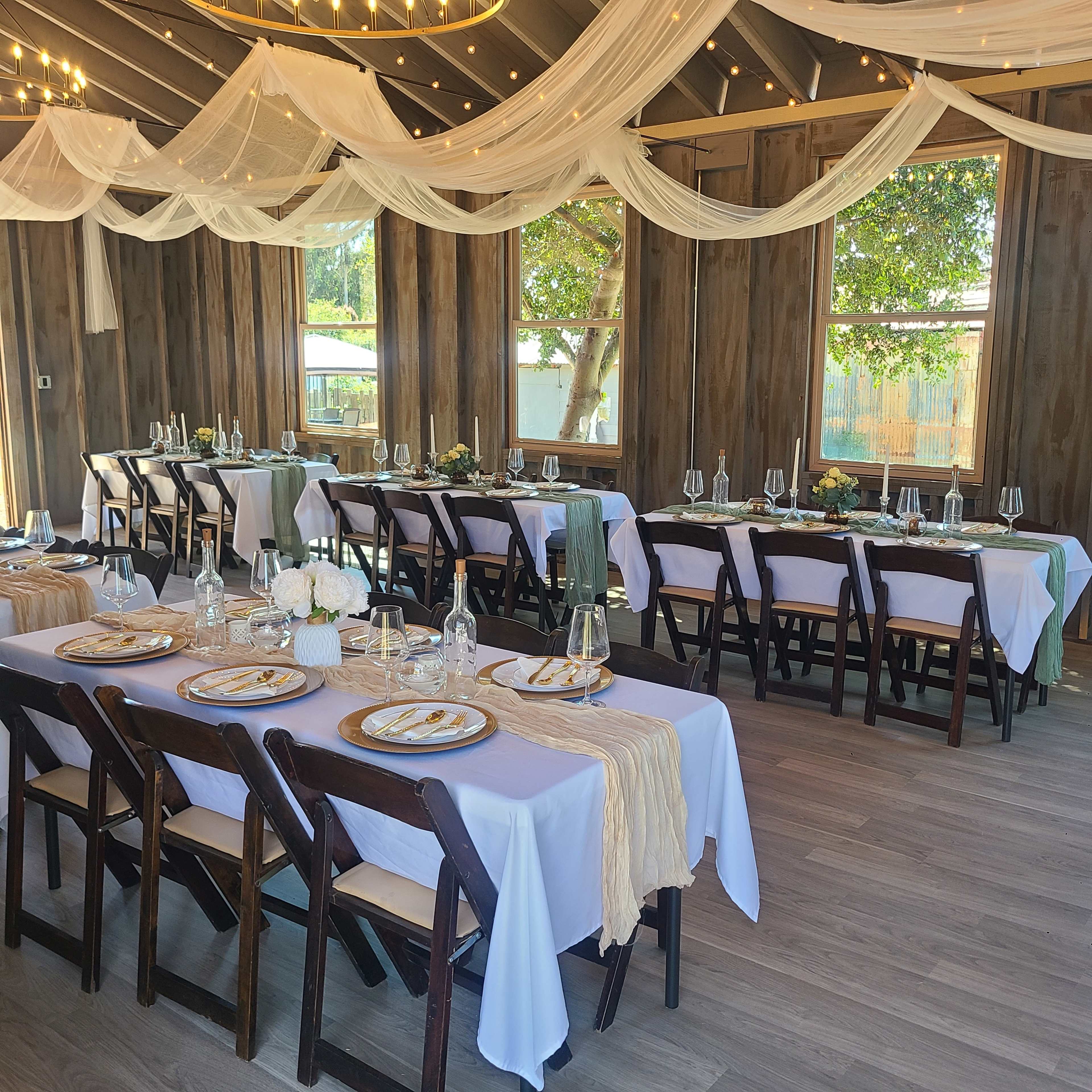 The image shows a decorated indoor venue with two long tables set for a meal, featuring white tablecloths, elegant place settings, and overhead drapery with string lights.