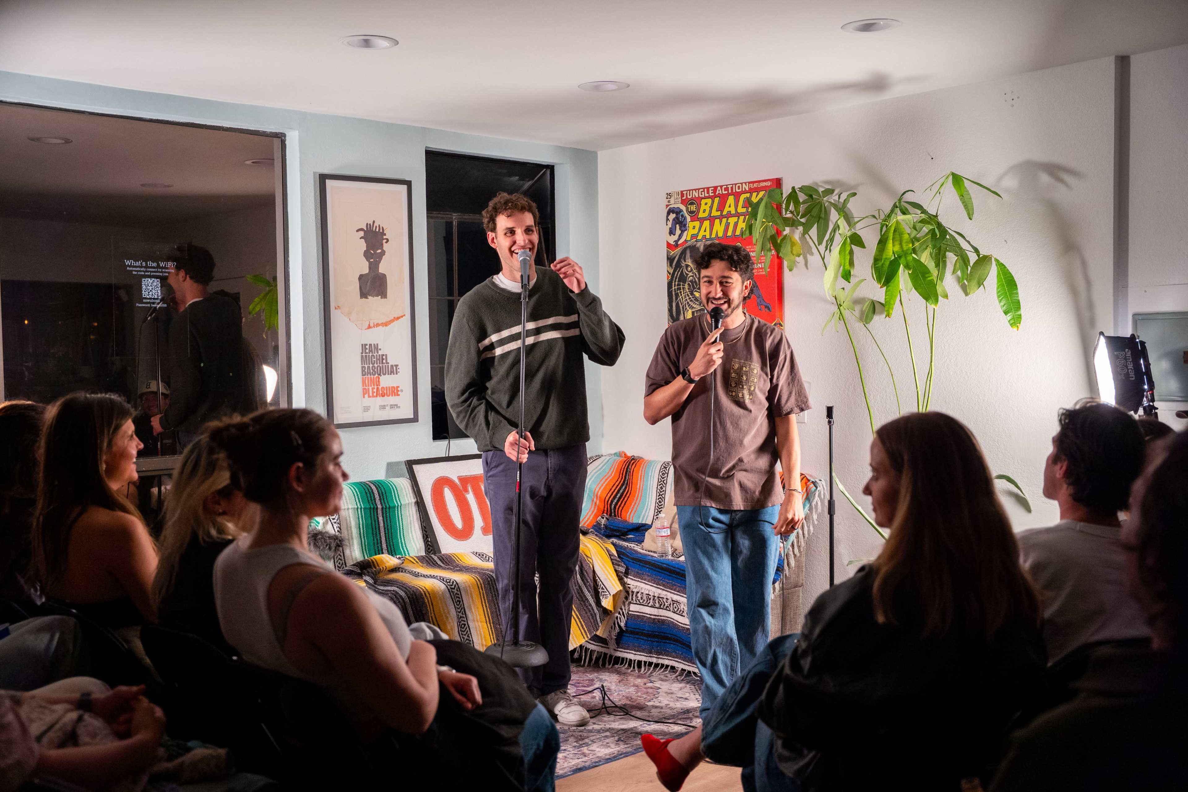 Two comedians perform stand-up in a cozy room with a small audience seated in front of them, surrounded by colorful decor and plants.