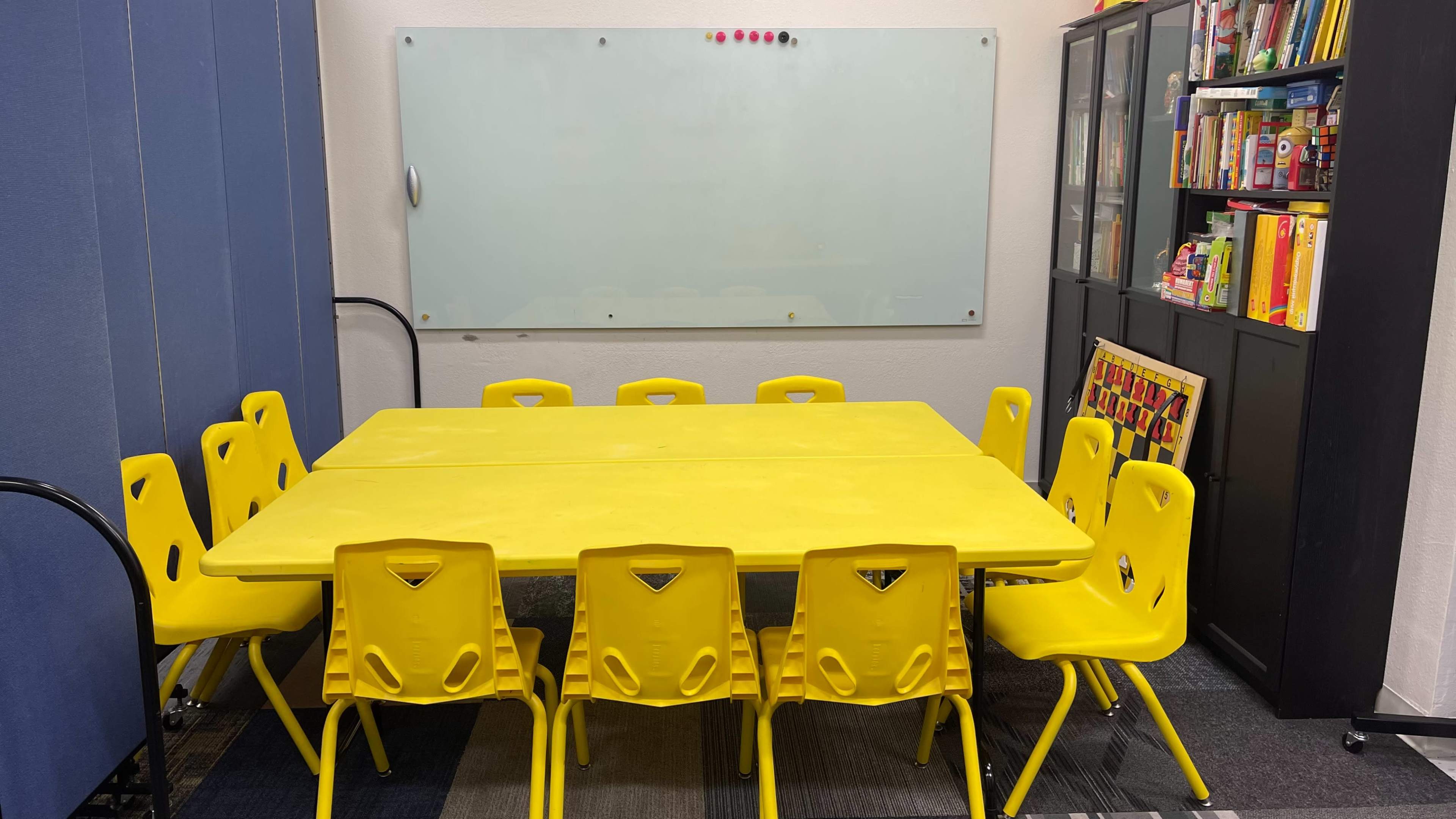 A large yellow table surrounded by twelve yellow chairs is positioned in a room with a whiteboard and a bookshelf filled with various items.