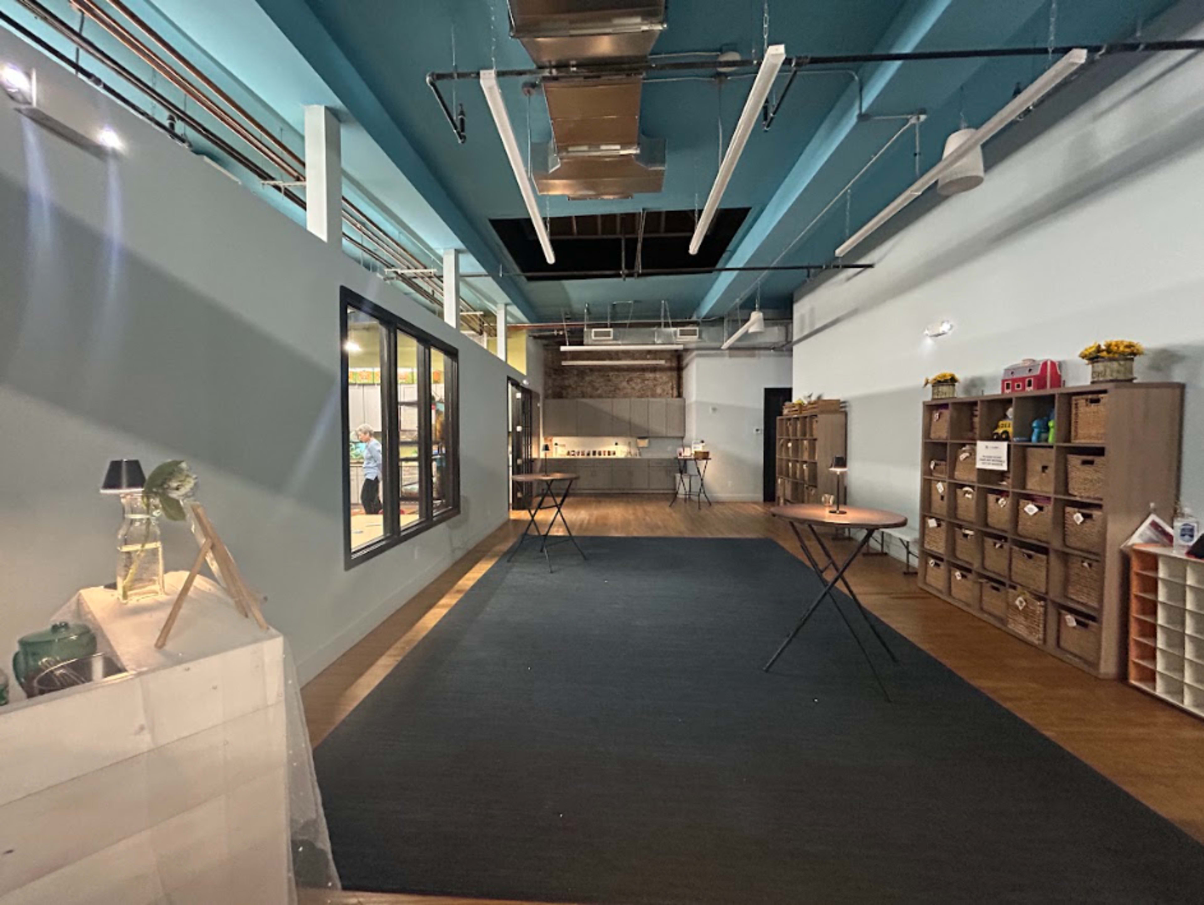 The image shows an open, well-lit interior space with wooden flooring, featuring tables, storage boxes, and a large window along one wall.